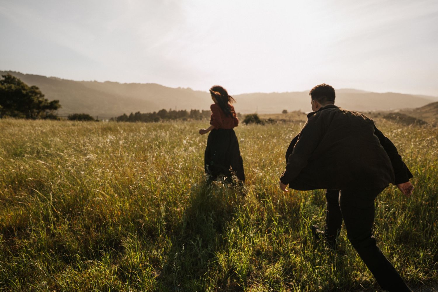 Couple running through golden sunlit meadow with rolling hills at sunset.Photography by The Y.K. Studio.