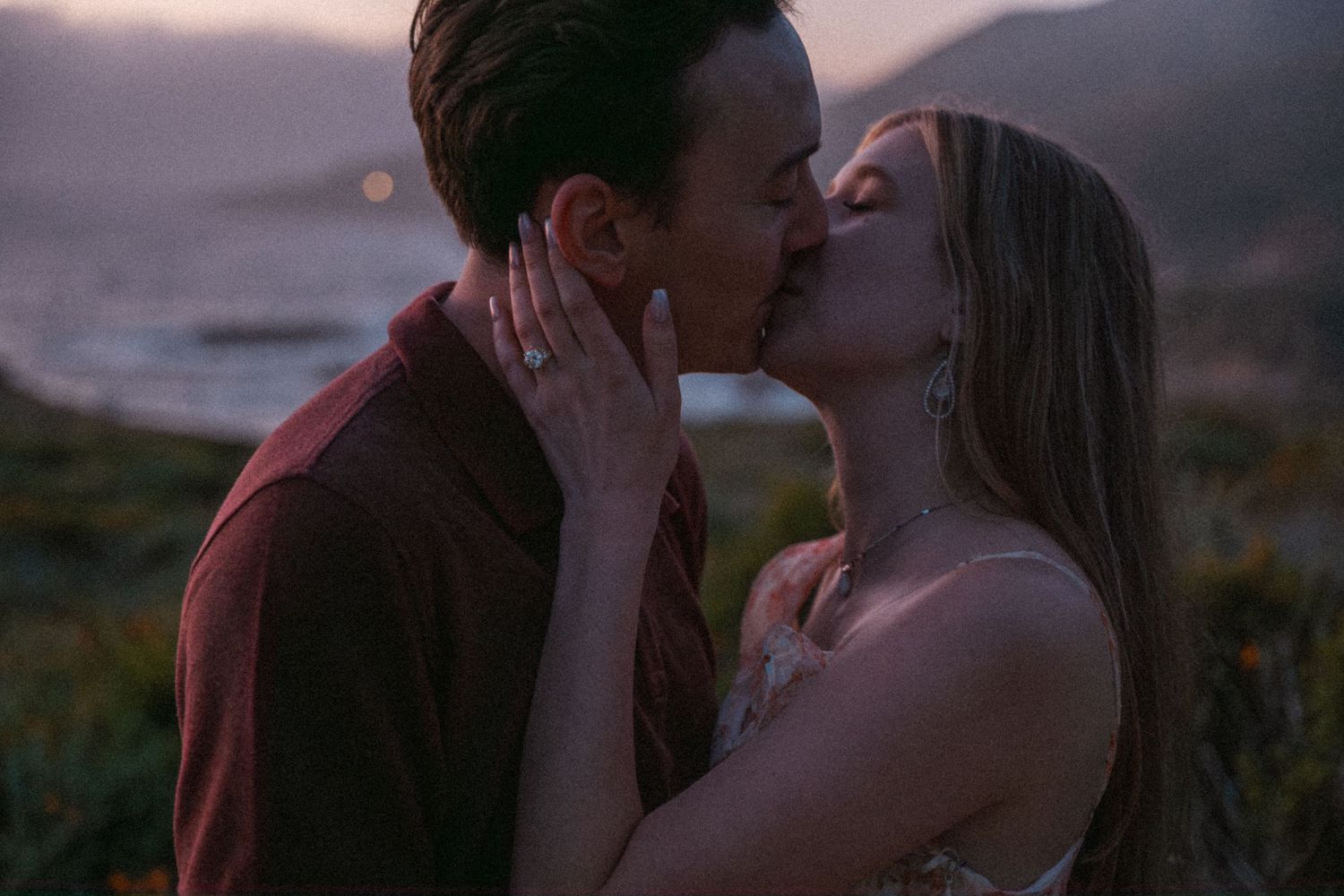 Couple sharing a romantic kiss at sunset with ocean and hills in the background.