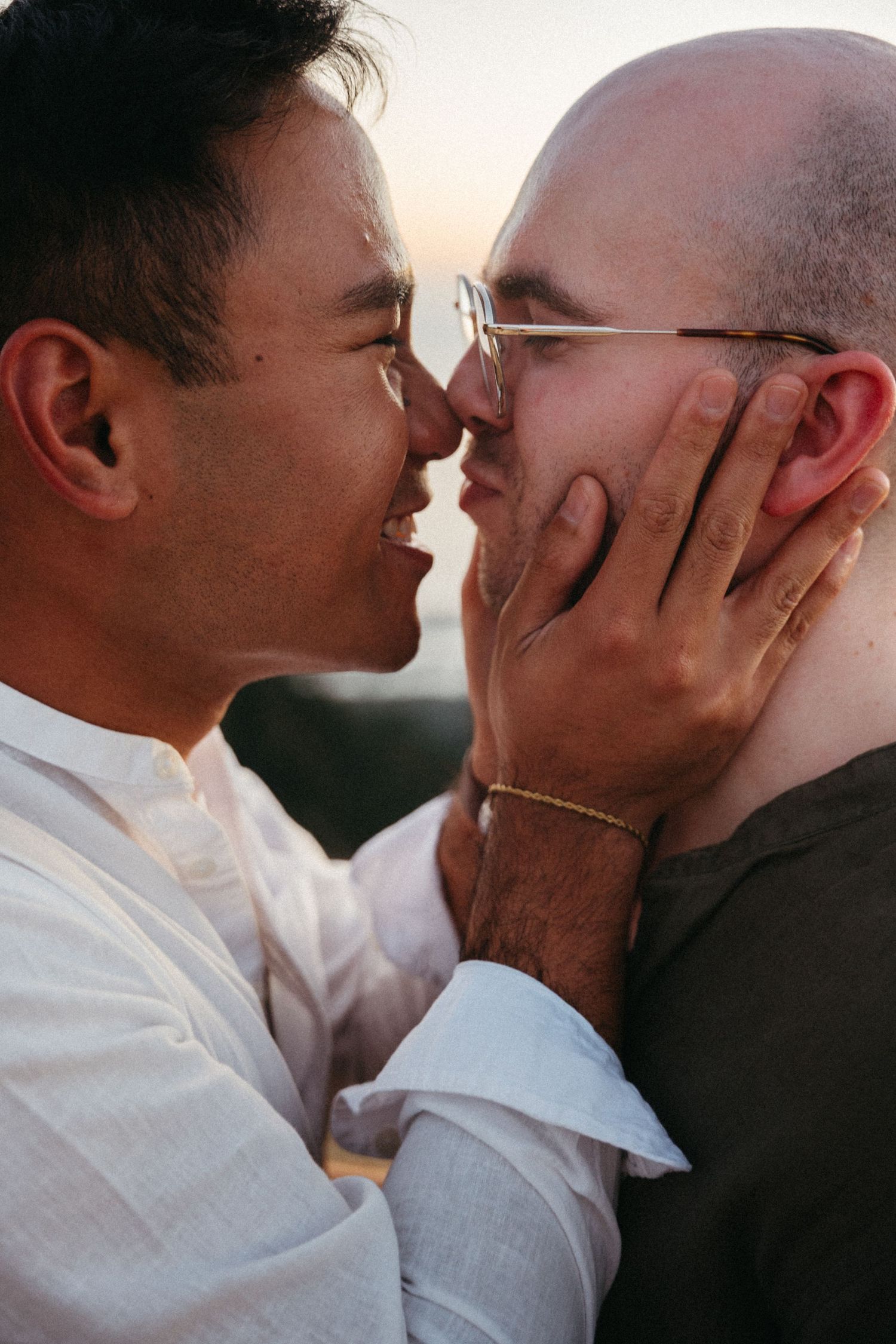 Two men share a tender romantic moment, one gently cupping the others face as they lean in for a kiss at sunset.