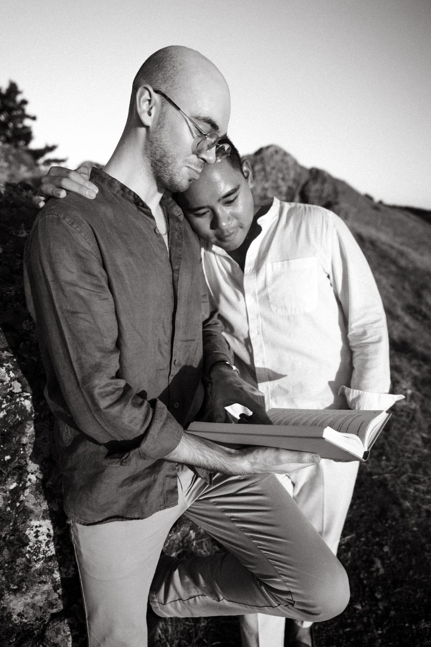 Two men share an intimate moment reading a book together outdoors in black and white photo.