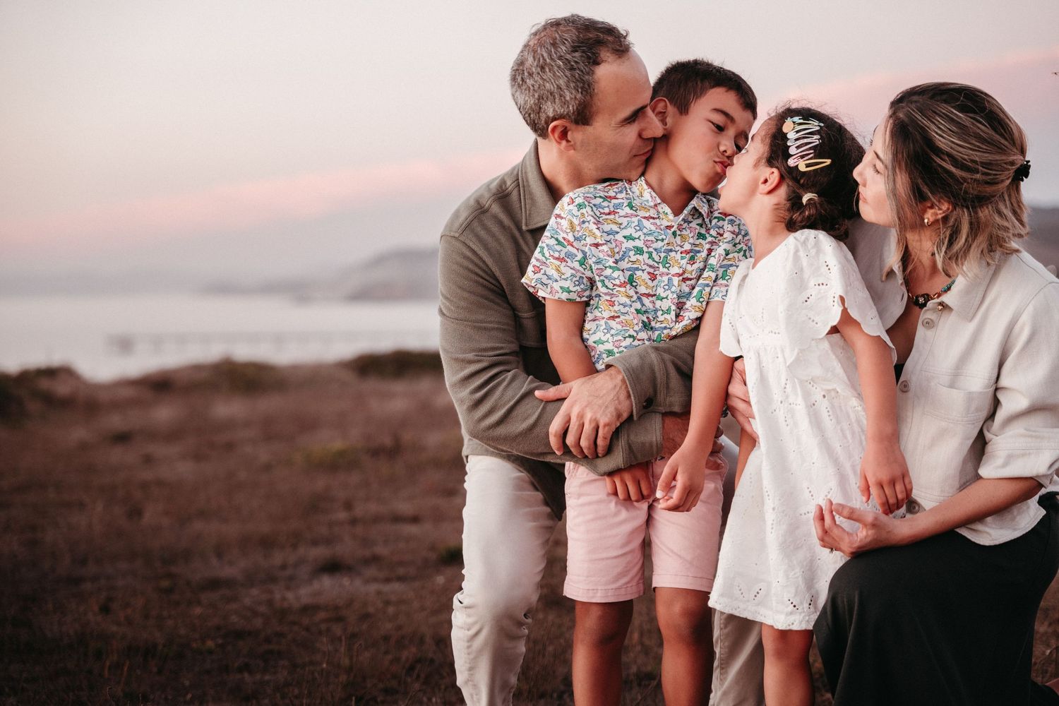 Family of four sharing kisses at sunset on a coastal landscape, parents kissing children in a tender outdoor moment.