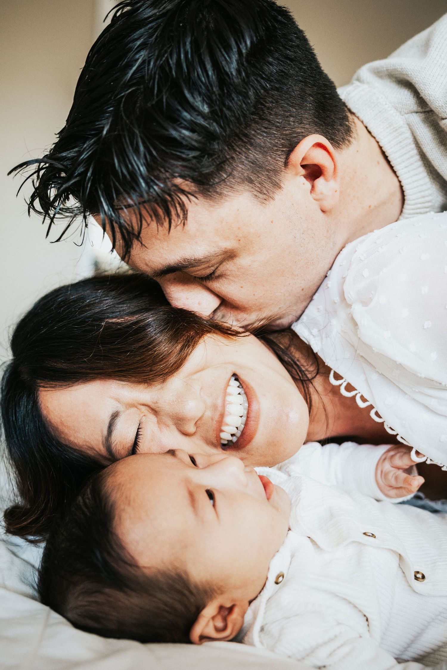 A man kisses a smiling woman on the forehead while they cuddle with their baby together. At home family session, Walnut Creek