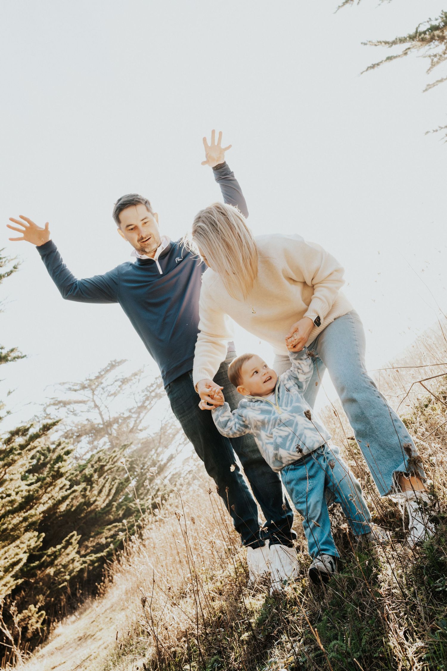 A joyful family of three plays outdoors, with parents raising their arms while holding their toddler on a sunny day. Mori Point, Pacifica.