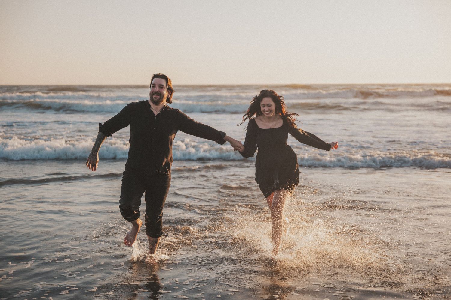 A joyful couple runs hand-in-hand through ocean waves on a beach at sunset, splashing water around them.