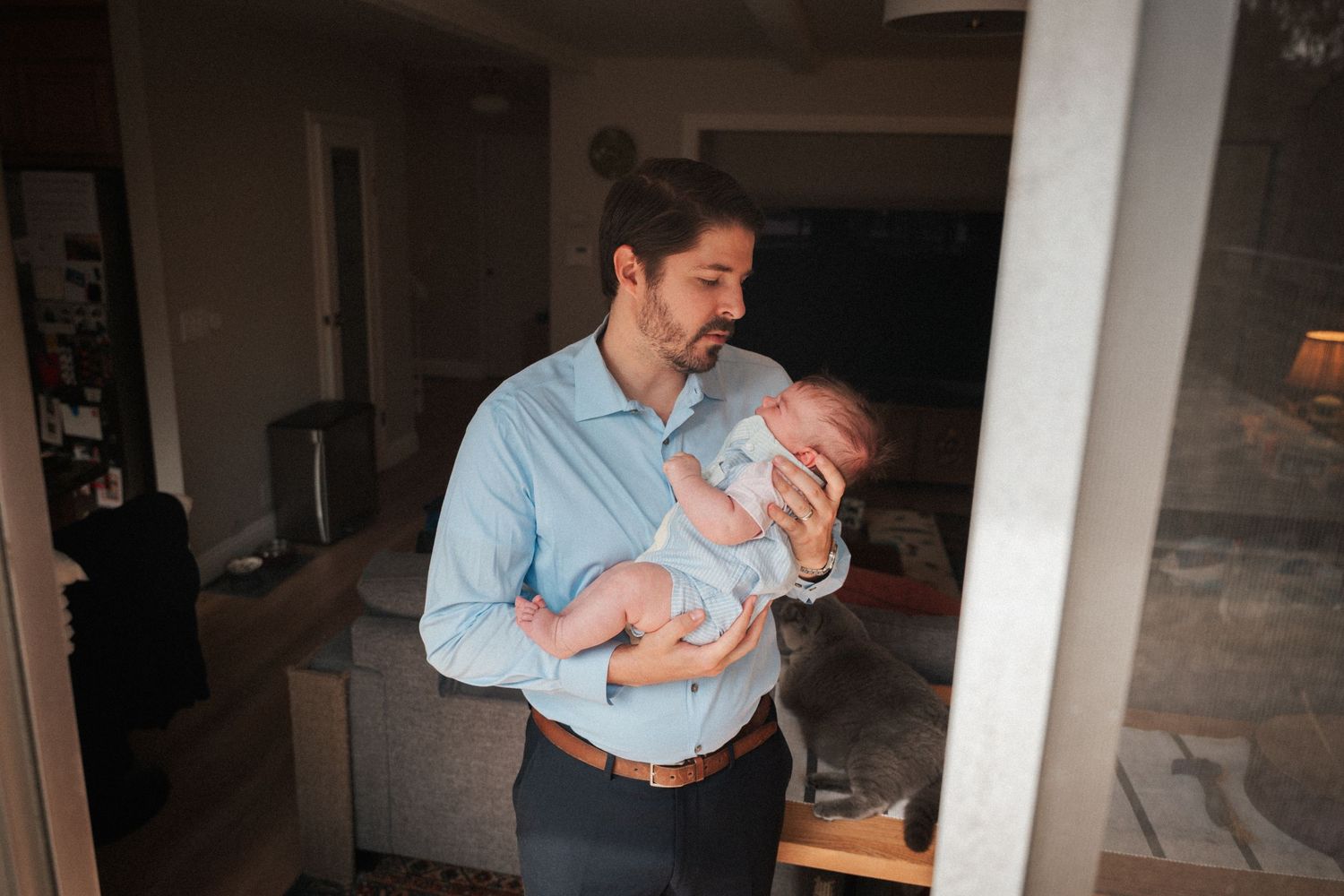 A man in a blue shirt tenderly holds a sleeping newborn baby inside a cozy home.