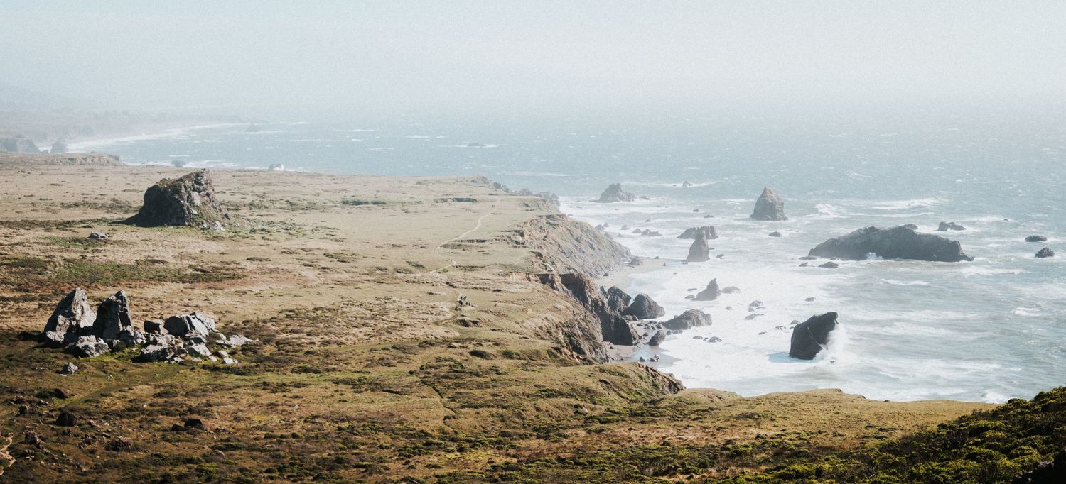 Couple sitting on a scenic coastal bluff overlooking misty ocean waves and rocky sea stacks in the distance.