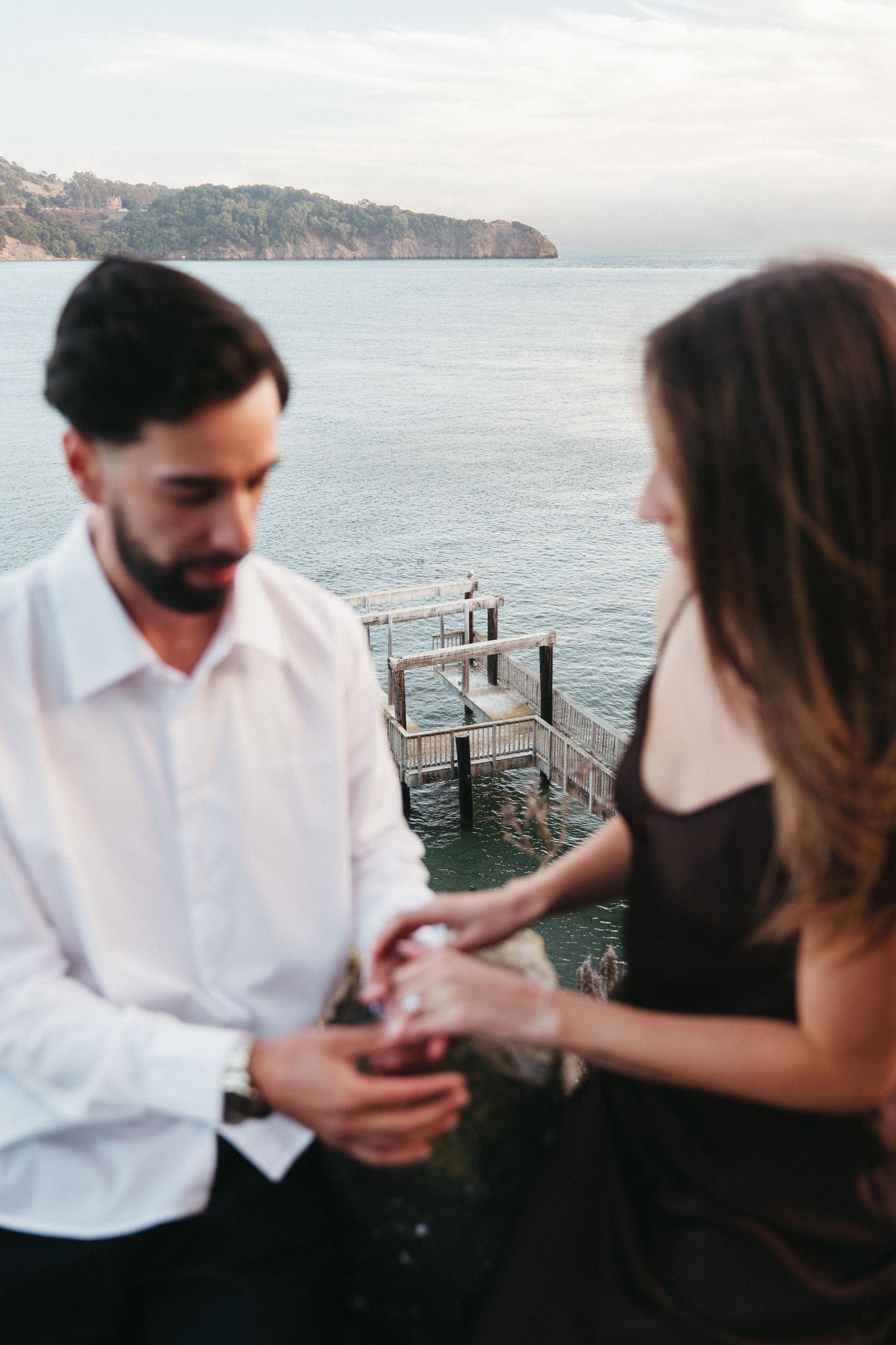 Couple sharing a romantic moment by the ocean, man in white shirt and woman in black dress holding hands near a pier.