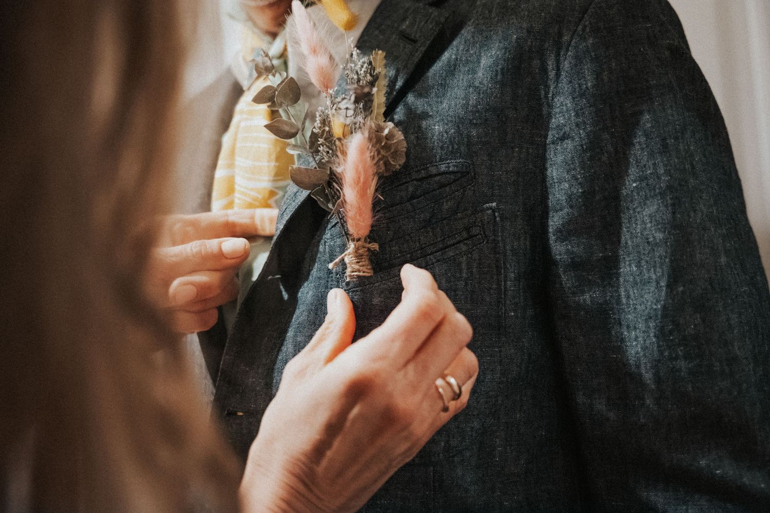 Close-up of hands pinning a dried floral boutonniere onto a dark tweed suit jacket.