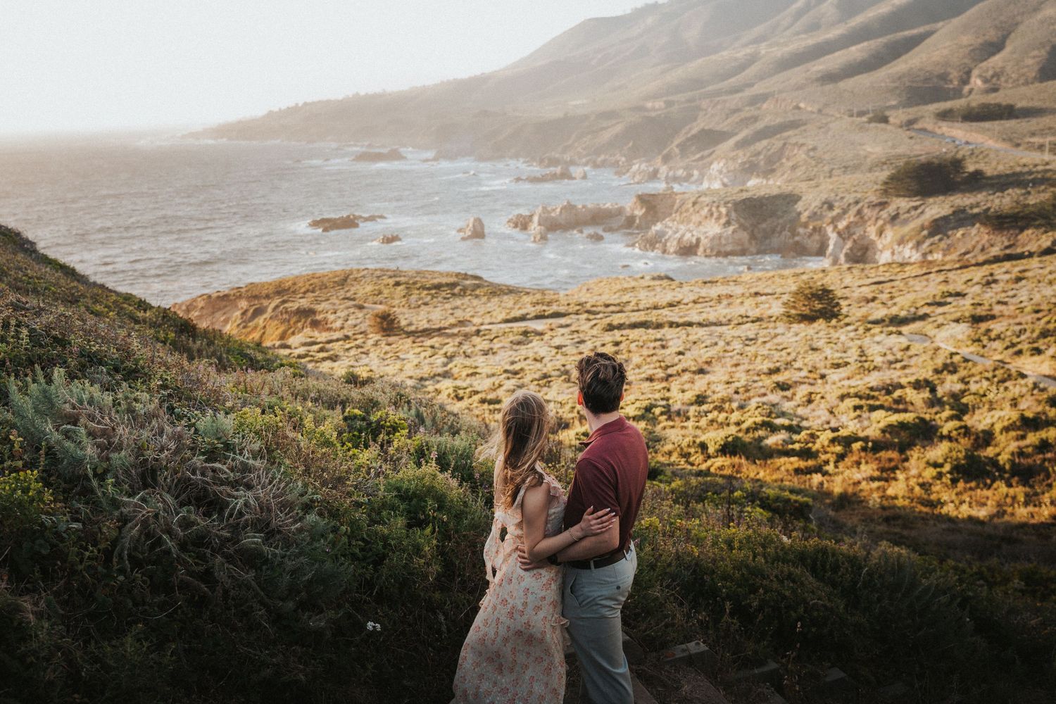 Couple embracing while overlooking a stunning coastal landscape with rocky cliffs and ocean views at golden hour.