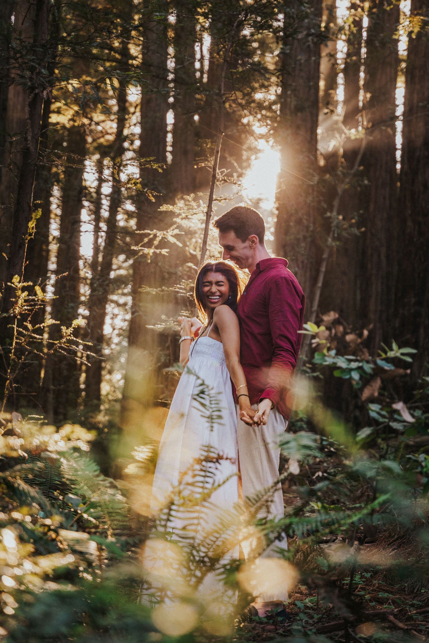 Couple embracing in sunlit forest, golden hour light streaming through tall trees, woman in white dress laughing joyfully.