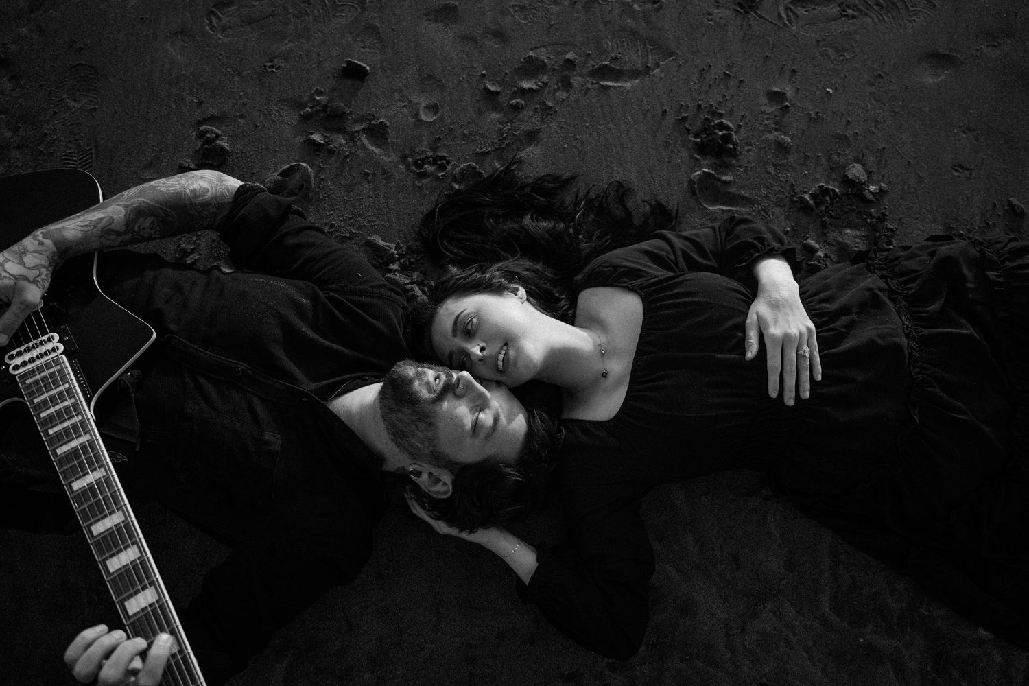 Black and white photo of two people lying on dark ground, one holding an acoustic guitar, heads together.