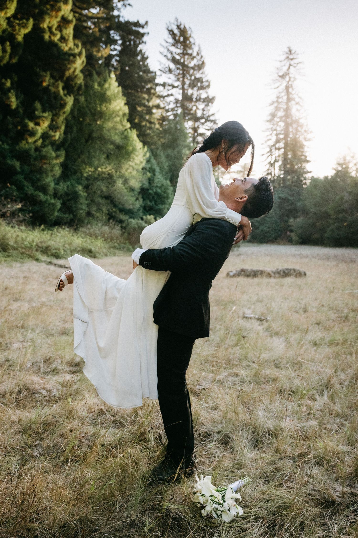 Groom lifting bride in white dress outdoors in a forest meadow during golden hour wedding photo.