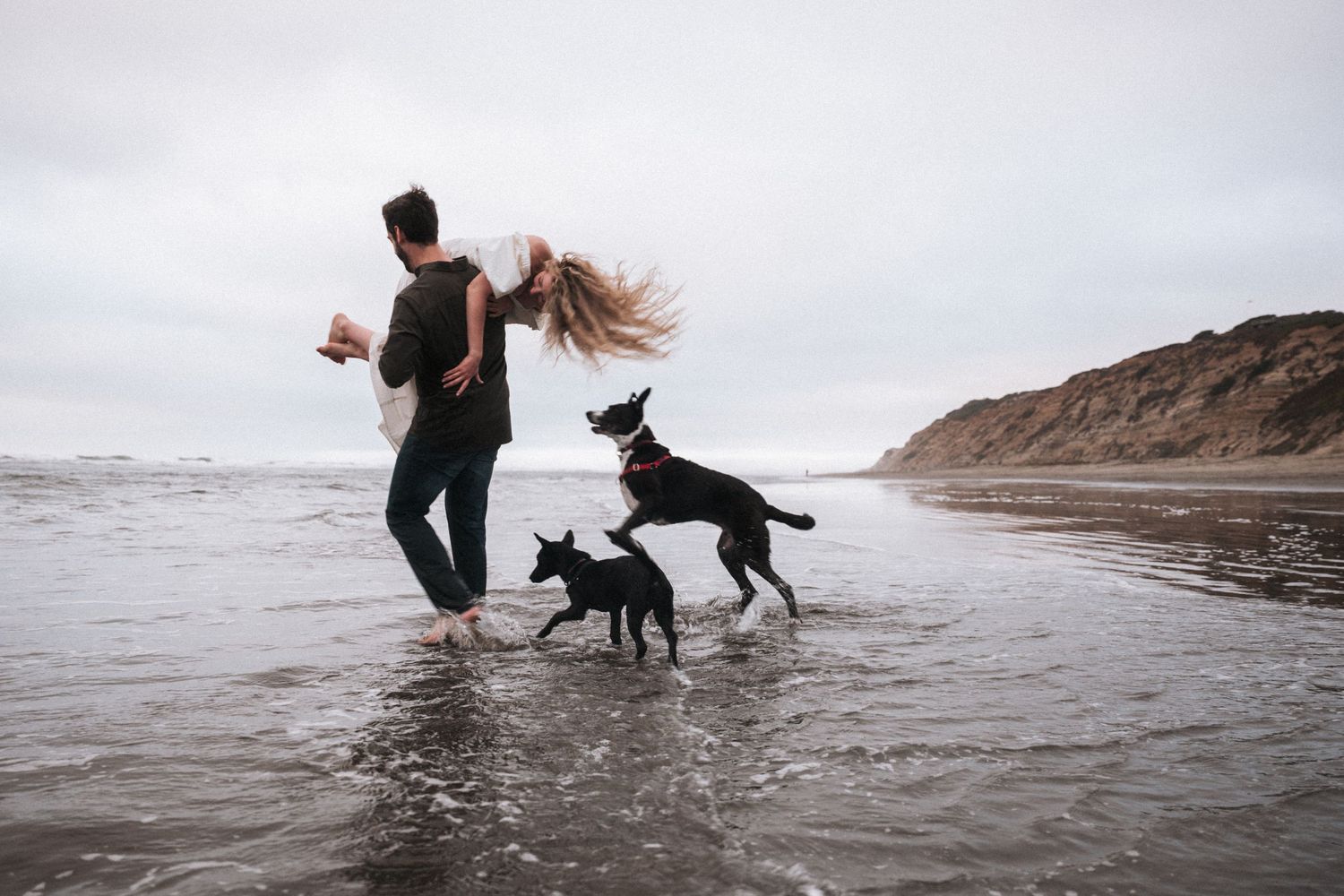 Couple playing with two black dogs on a sandy beach with ocean waves and cloudy sky.