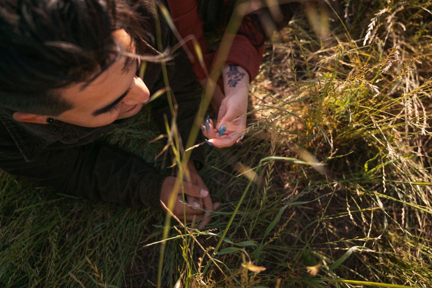 Person examining plants closely in a sunlit grassy field, with a colorful tattoo visible on their hand.Photography by The Y.K. Studio.