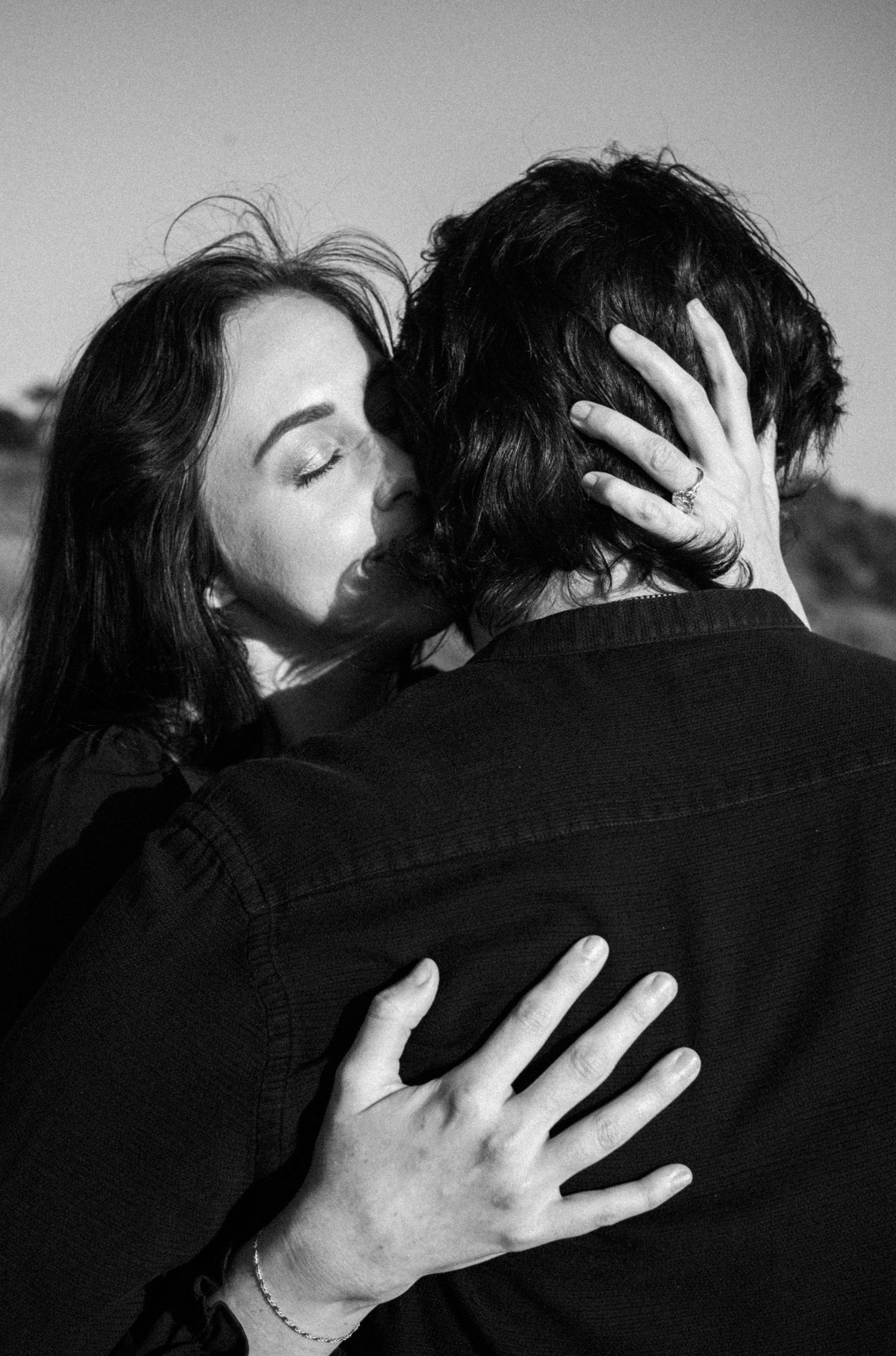 Woman with long dark hair embracing and kissing a man outdoors in black and white photo.