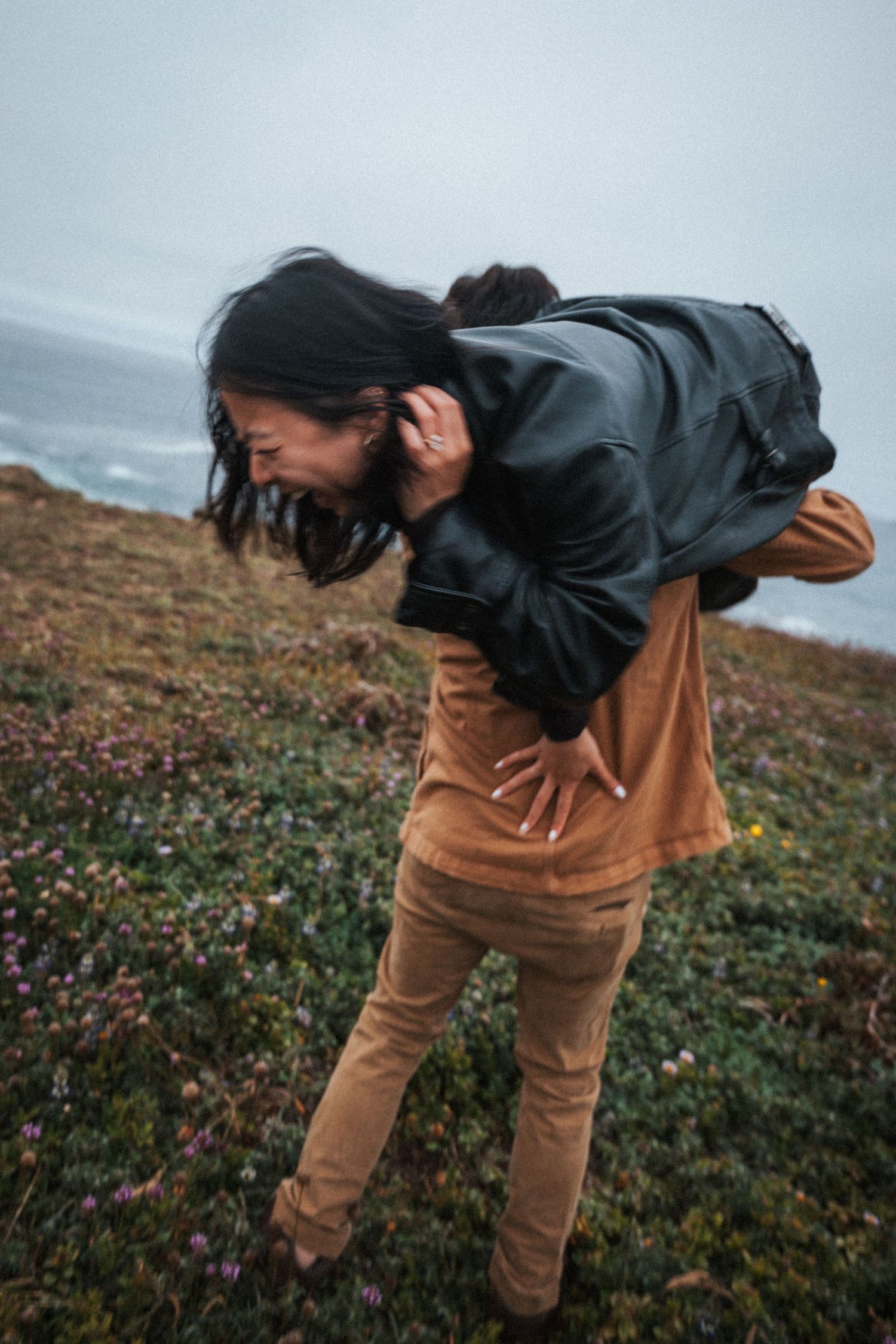 A woman in a leather jacket and tan pants laughs while being carried on a coastal hillside on a cloudy day.