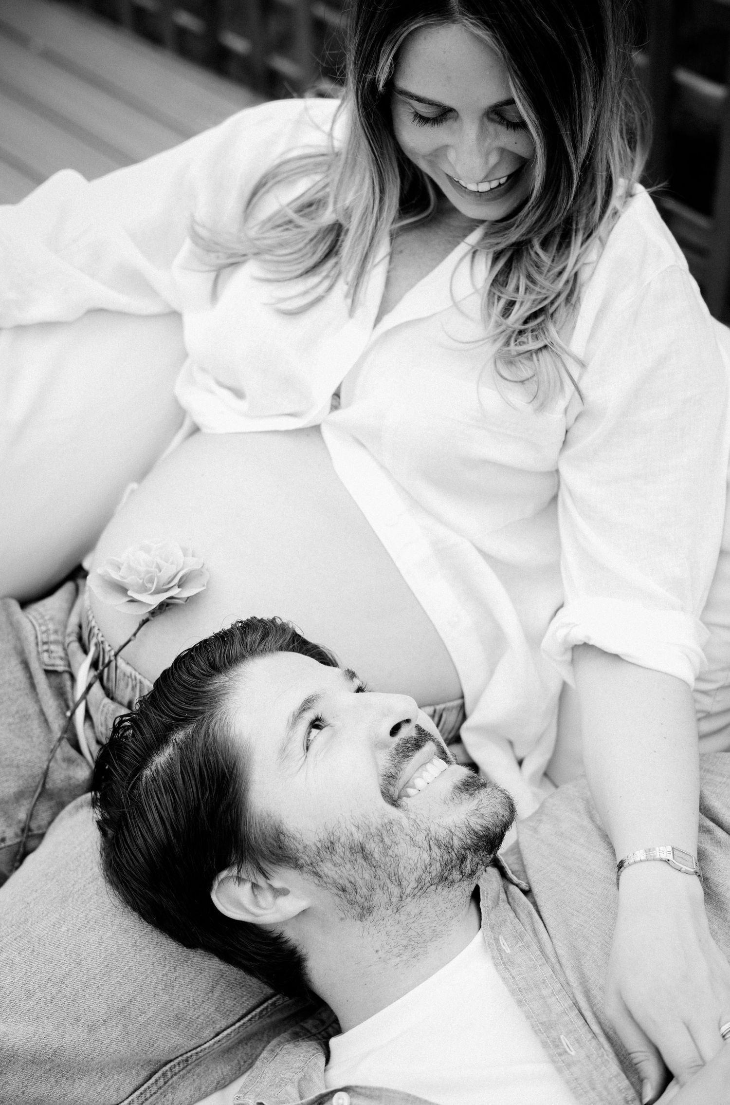 Black and white maternity photo of smiling couple, man lying with head near pregnant woman's belly outdoors.