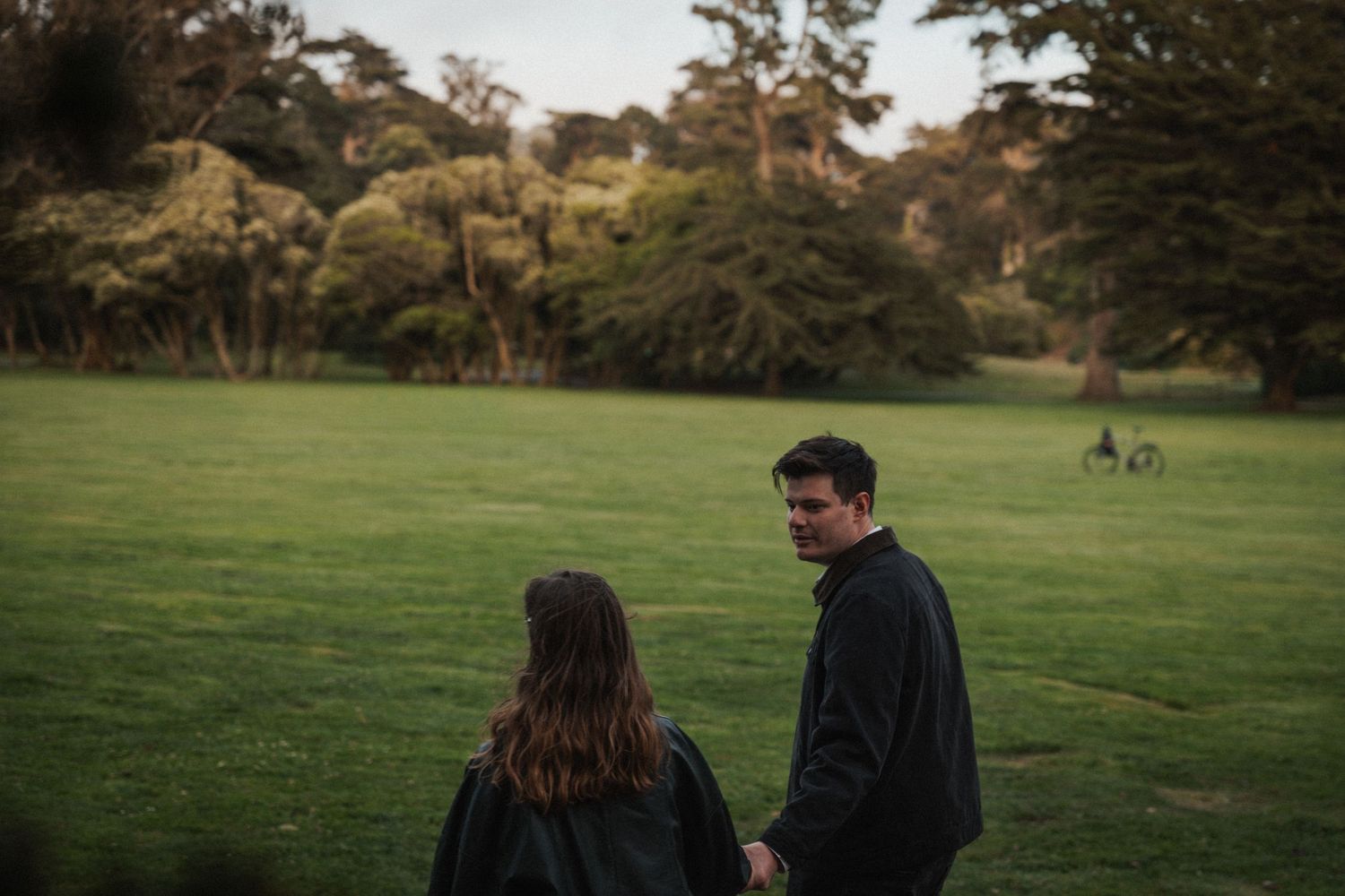 Couple holding hands walking through a lush green park surrounded by tall trees.