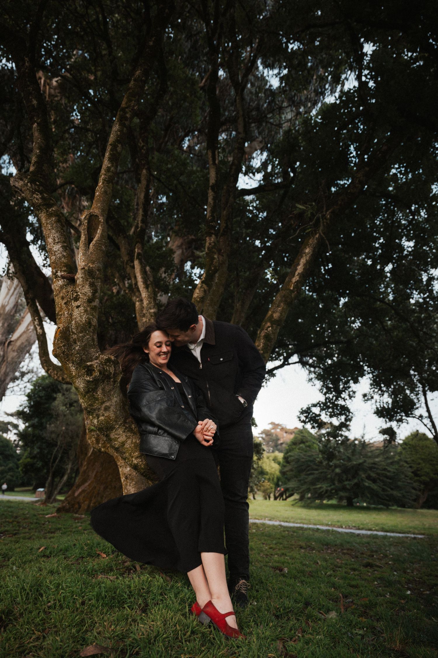 Couple laughing and leaning against a large moss-covered tree in a lush park setting.