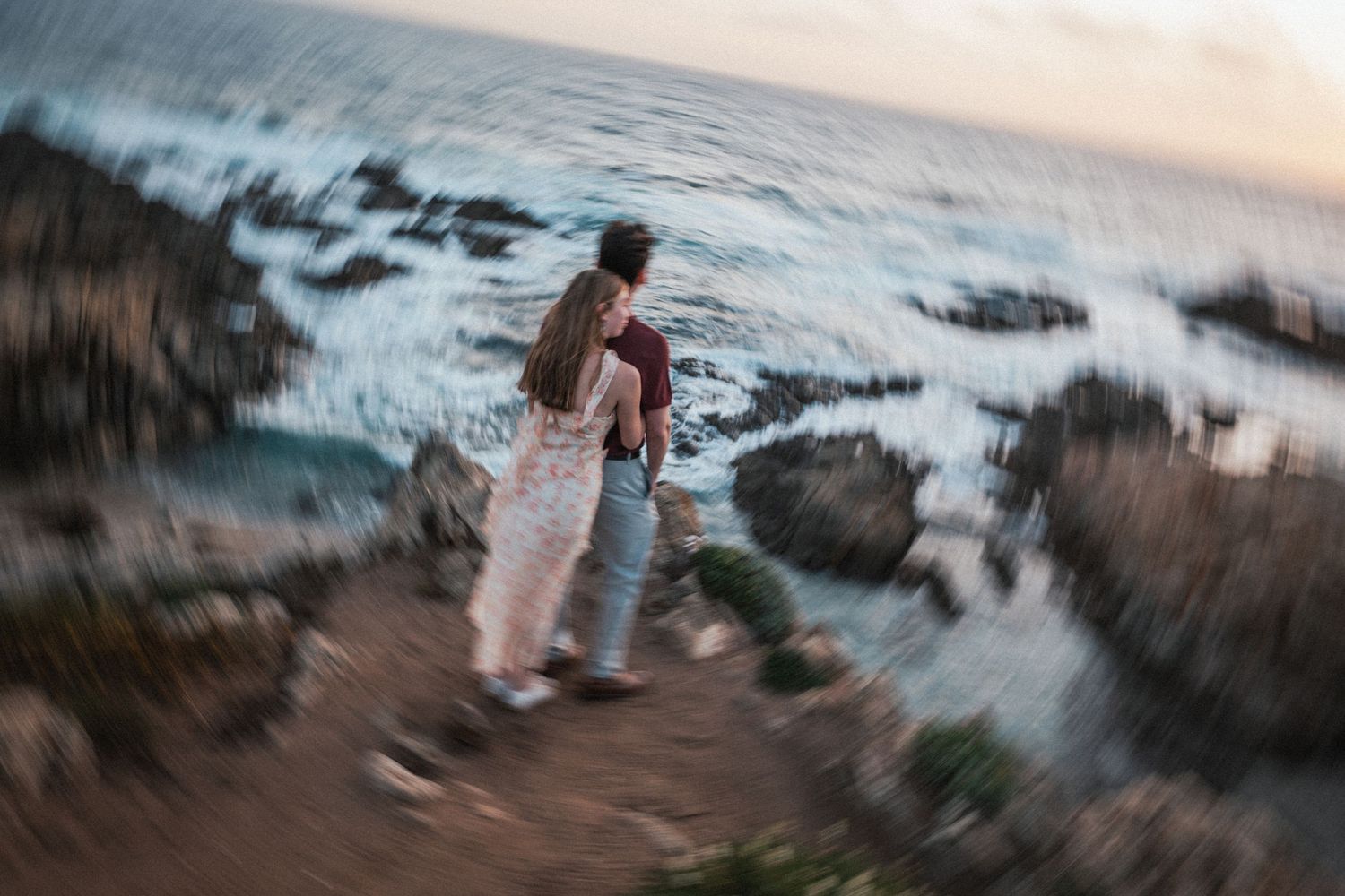 Couple standing together on rocky coastal cliffs above crashing ocean waves.