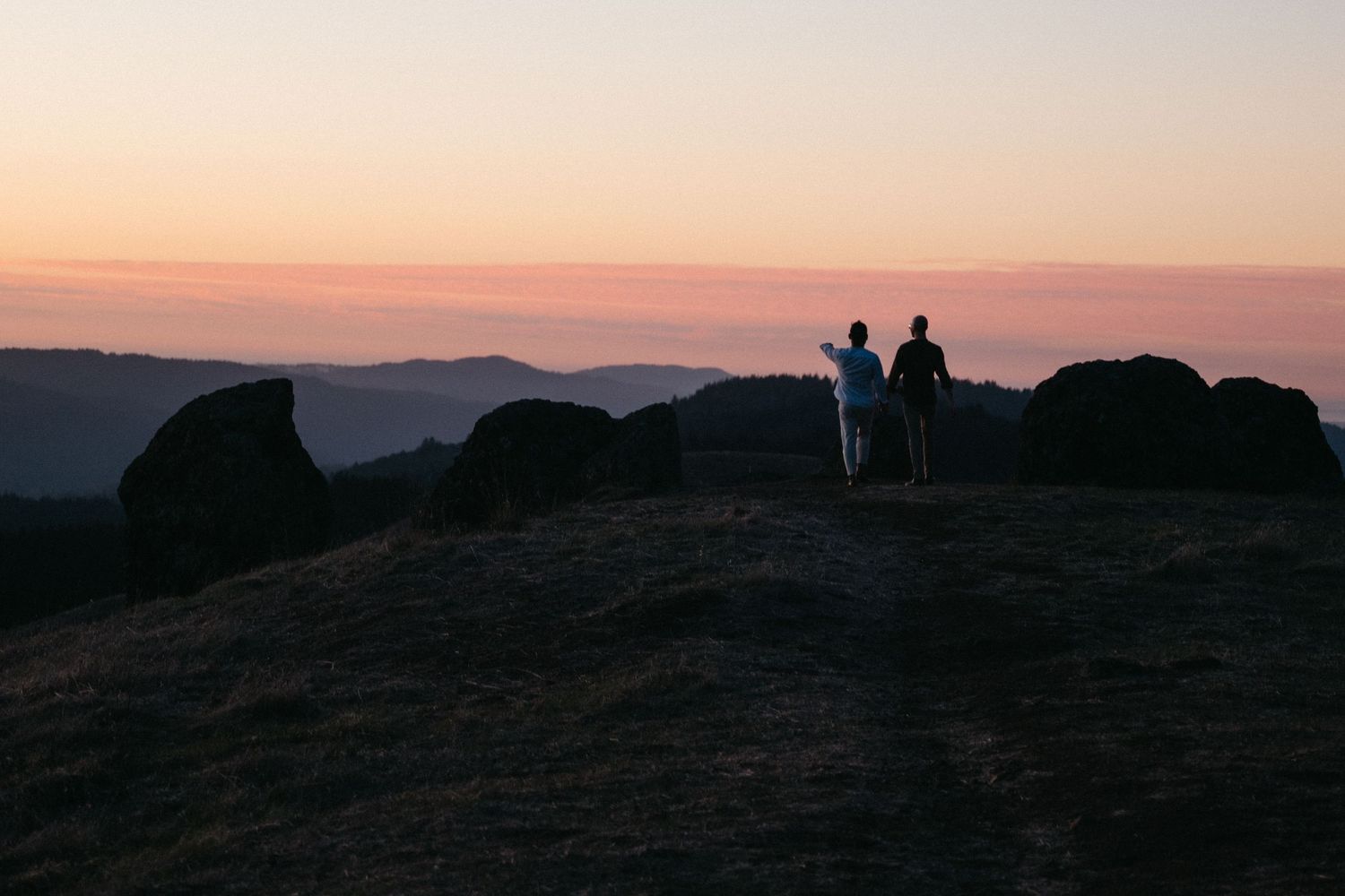 Two silhouetted figures stand on a rocky hilltop at sunset, overlooking a serene mountain landscape with a pink sky.