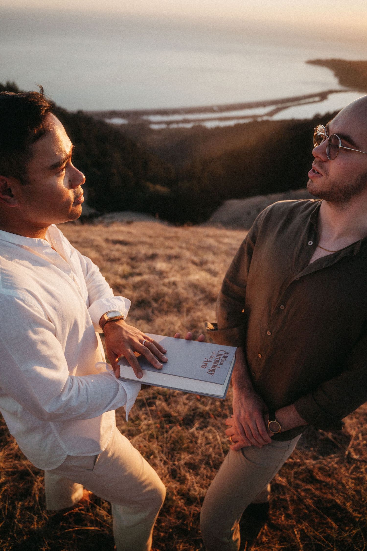 Two men exchange vows outdoors on a hillside at sunset during an intimate elopement ceremony.