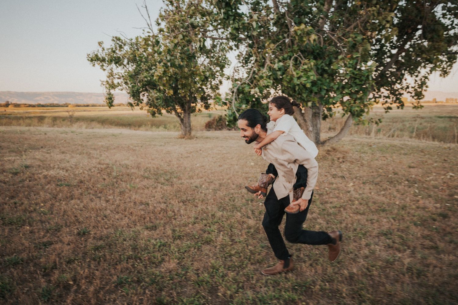A father gives his child a piggyback ride through a grassy field with trees in the background.