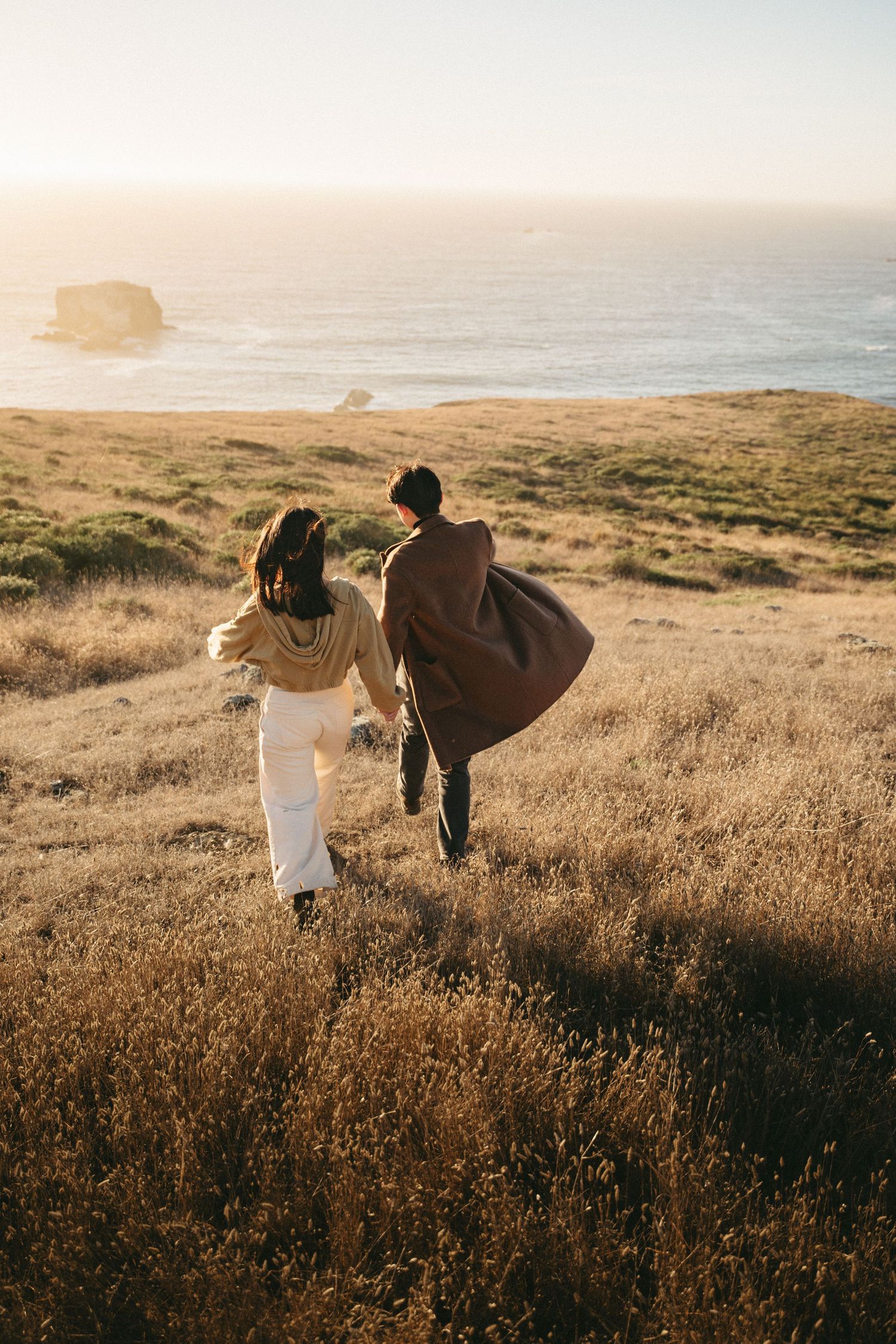 Couple in warm autumn clothing run hand in hand through golden coastal grass toward the ocean at sunset.