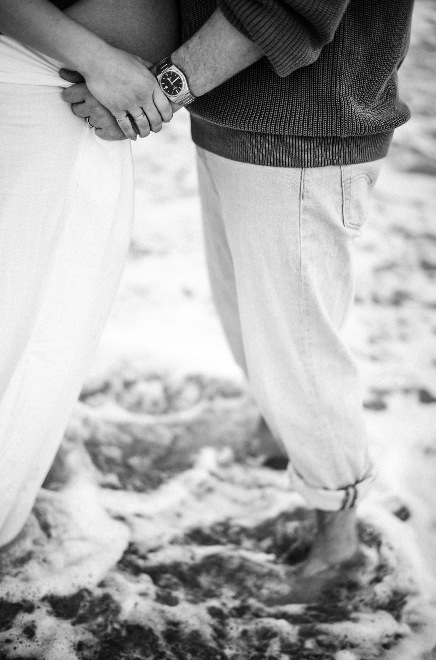 Black and white close-up of couple holding hands on sandy beach, man wearing watch and rolled jeans.