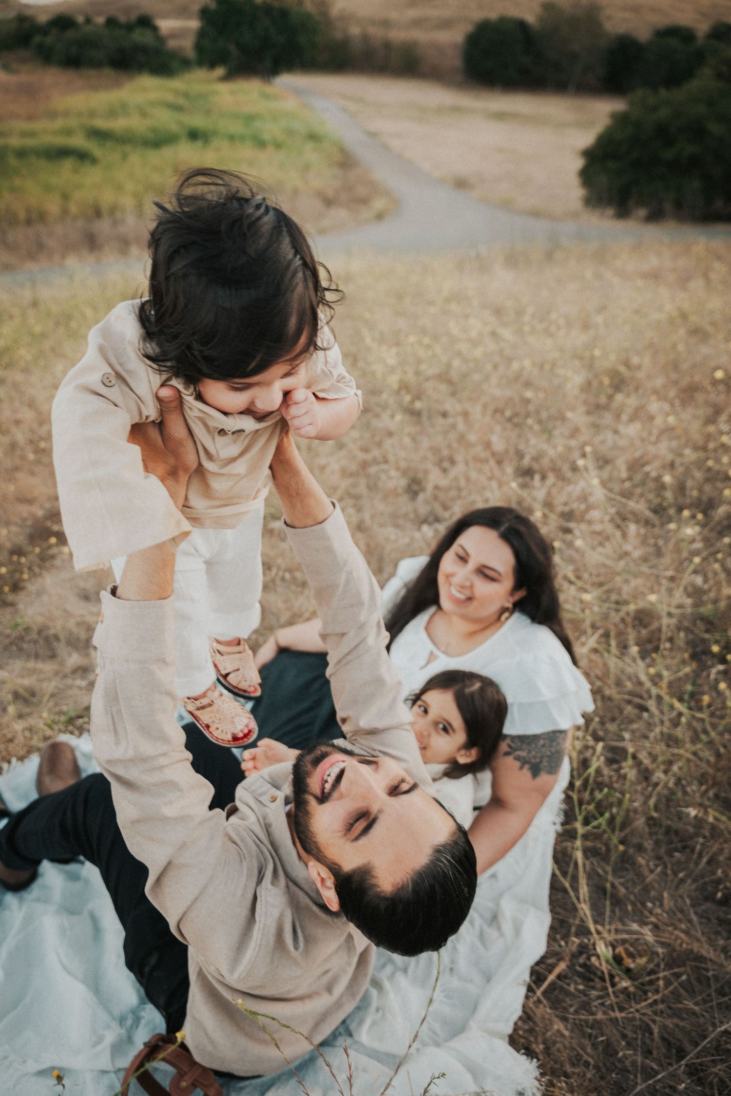 A joyful father lies in a field lifting his toddler overhead while mother and child smile nearby.