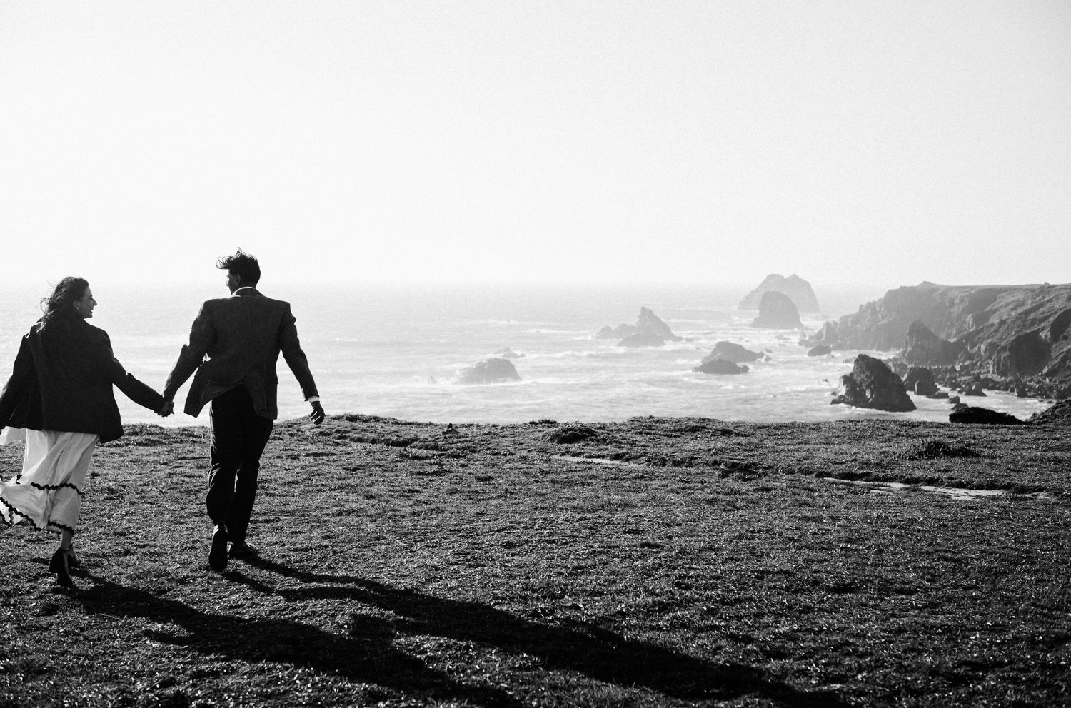 Black and white photo of a couple holding hands walking toward rocky ocean cliffs on a grassy coastal bluff.Photography by The Y.K. Studio.