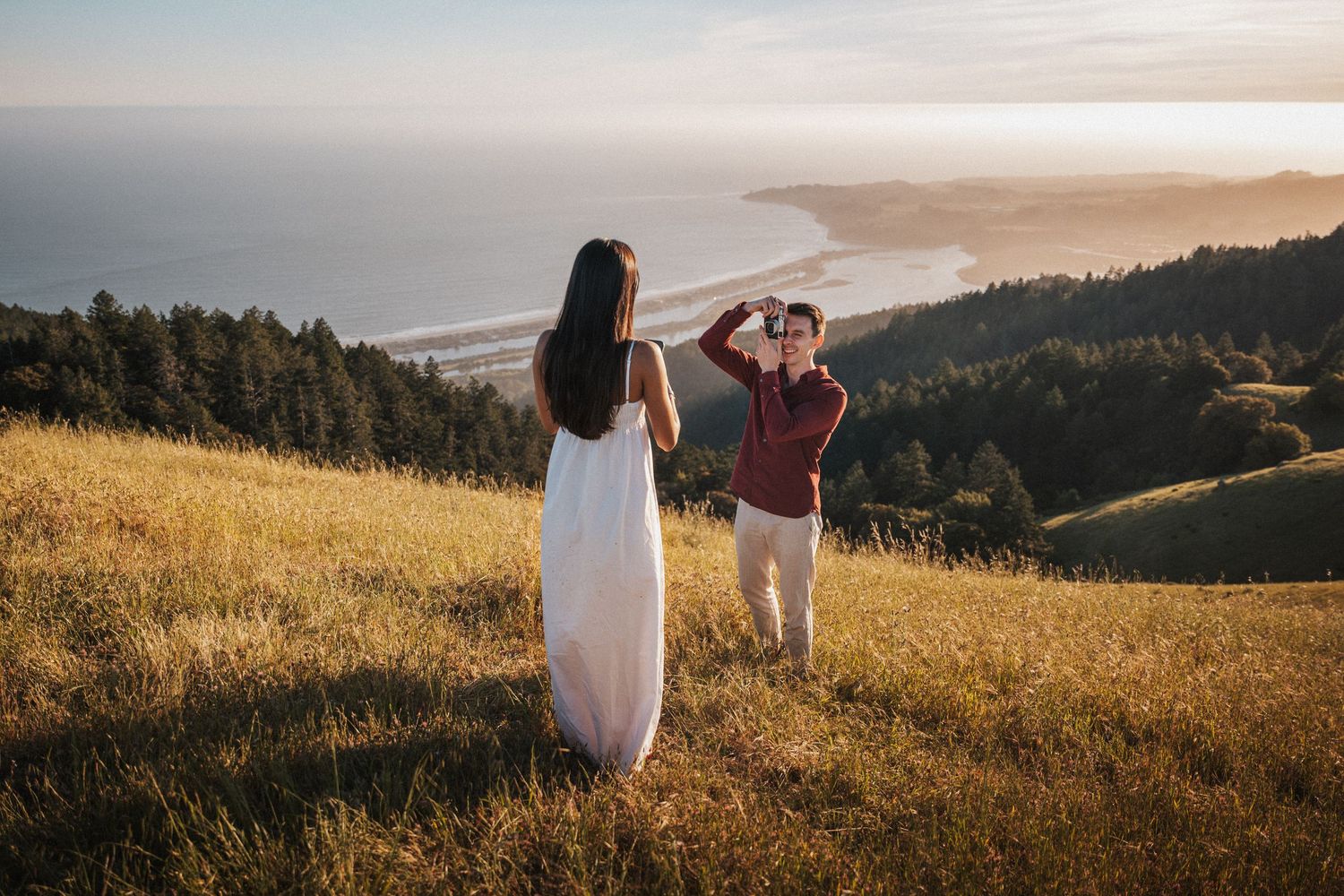 Couple on a golden hillside, woman in white dress poses as man photographs her with coastal ocean views behind them.