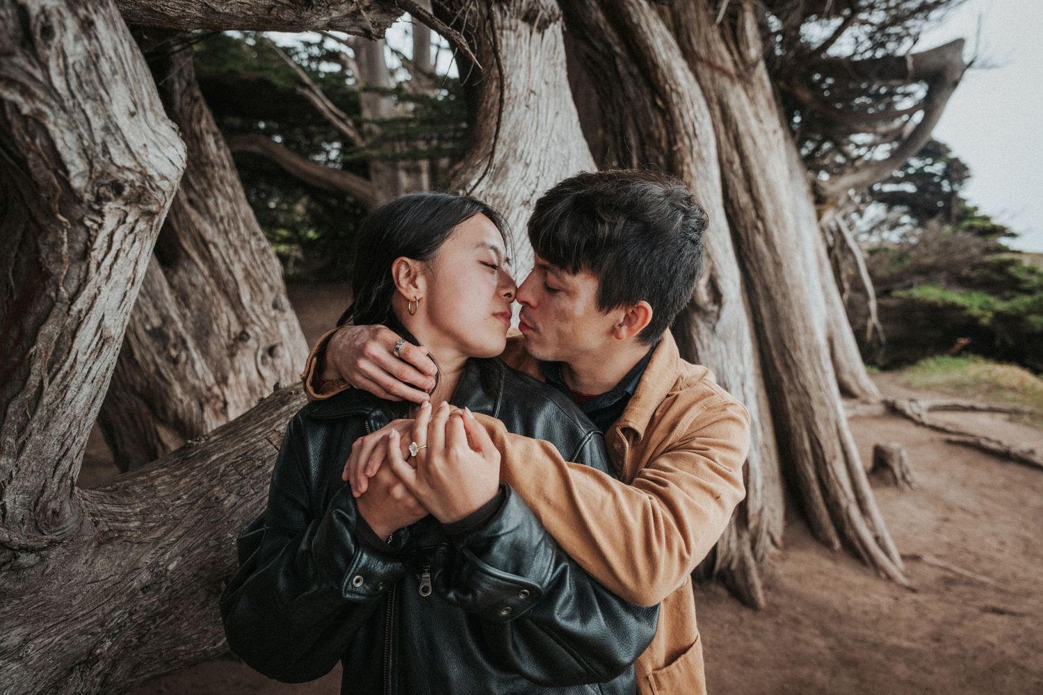 Couple sharing a tender kiss embrace near ancient twisted trees in a romantic outdoor setting.