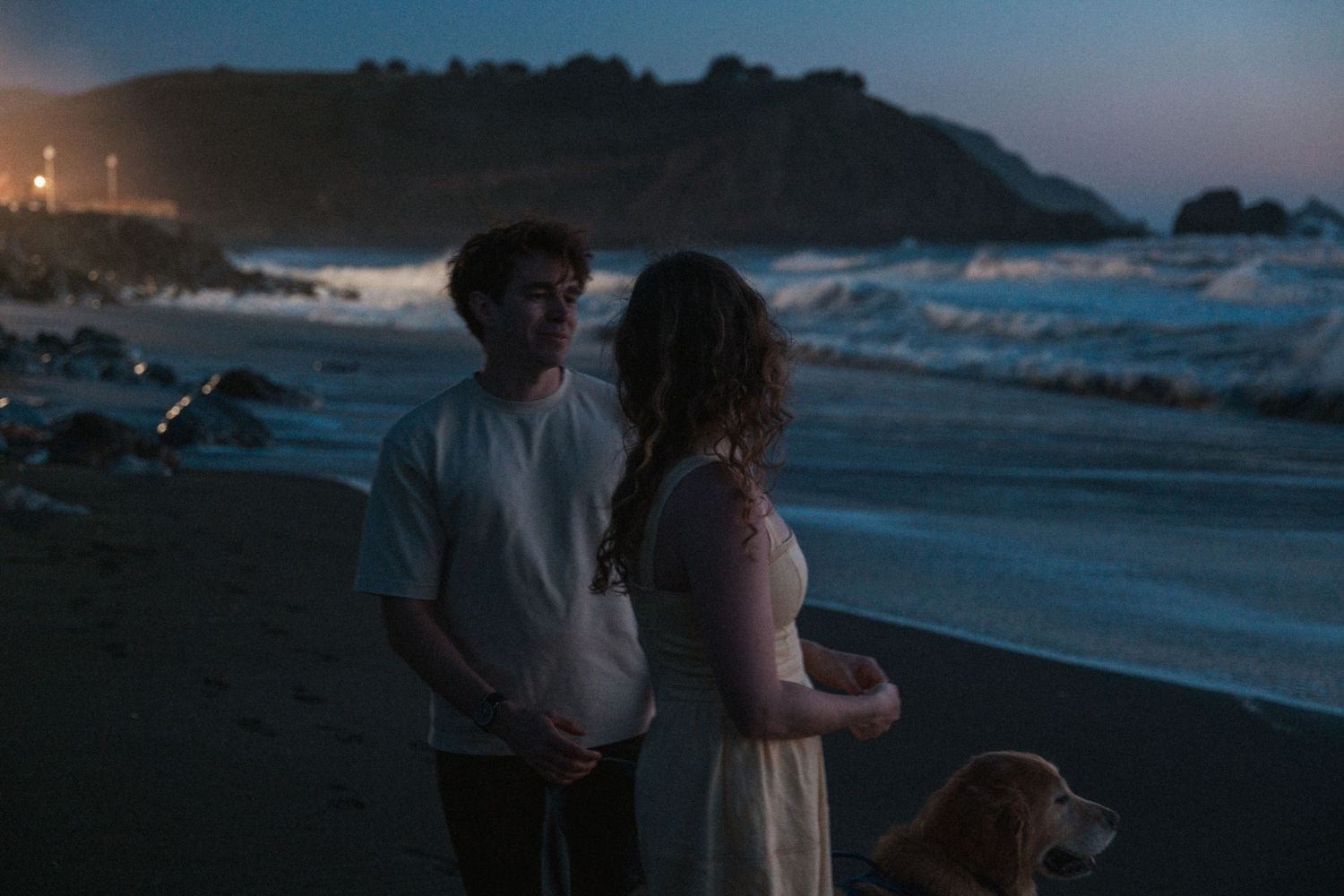 Romantic couple standing on a beach at dusk with their dog by the waves.