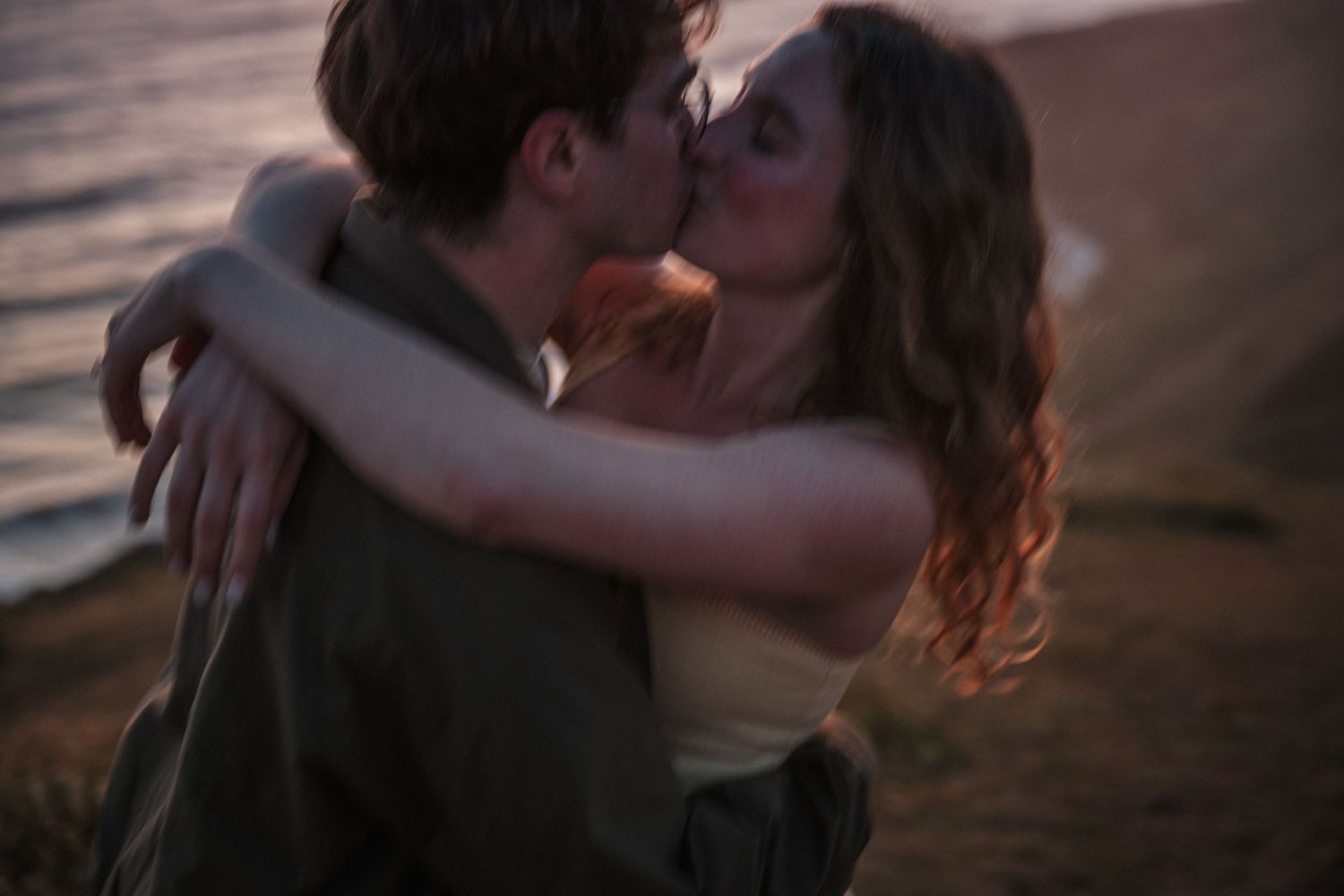 A couple shares a romantic kiss on a beach at sunset, embracing each other near the ocean shore. Photography by The Y.K. Studio.