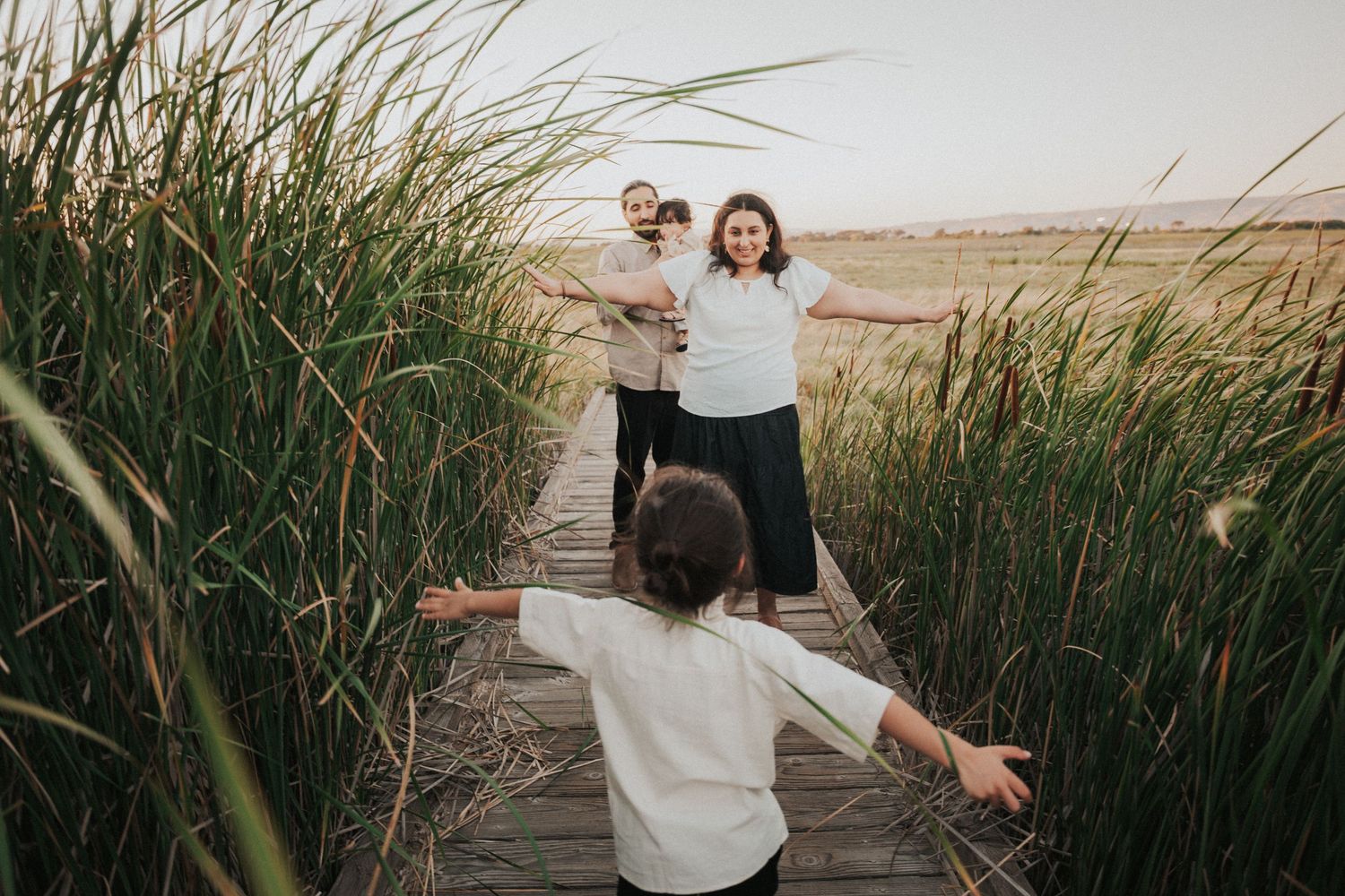 Child running with arms outstretched on wooden boardwalk toward family surrounded by tall green reeds at sunset.