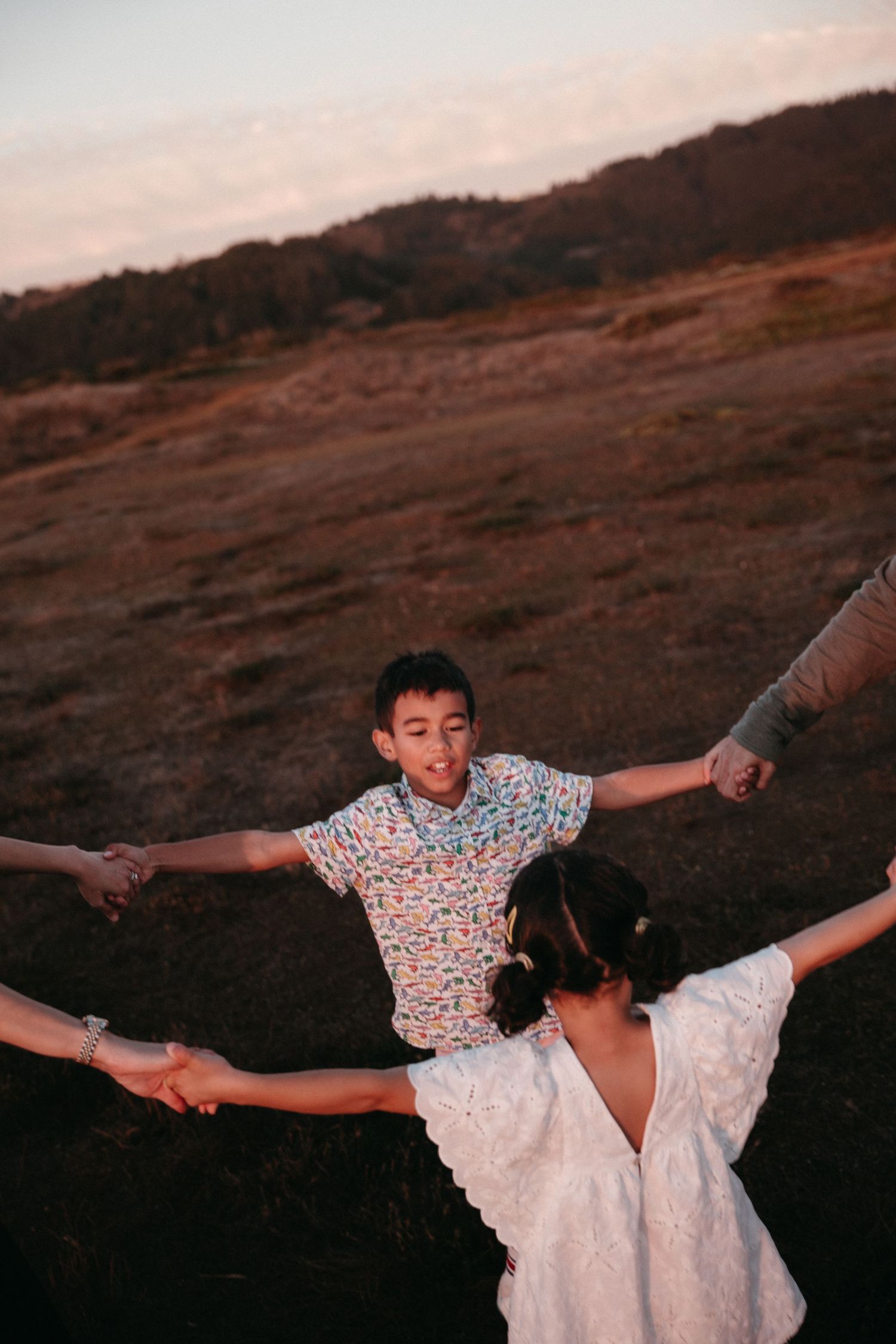 A boy in a colorful shirt holds hands with two people on a hillside at sunset.