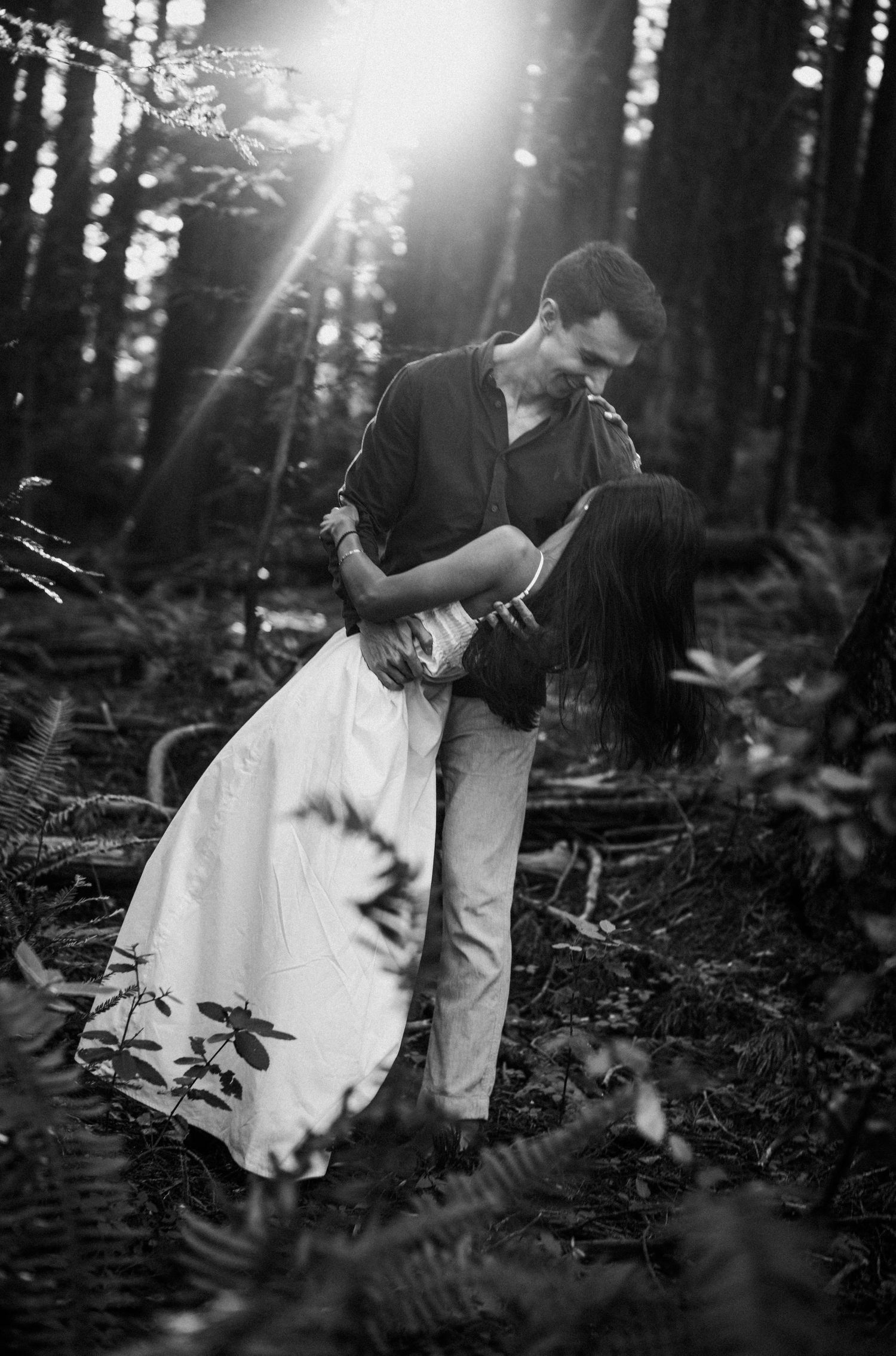 Black and white photo of a couple sharing a romantic kiss in a sunlit forest, woman in white dress.Photography by The Y.K. Studio.