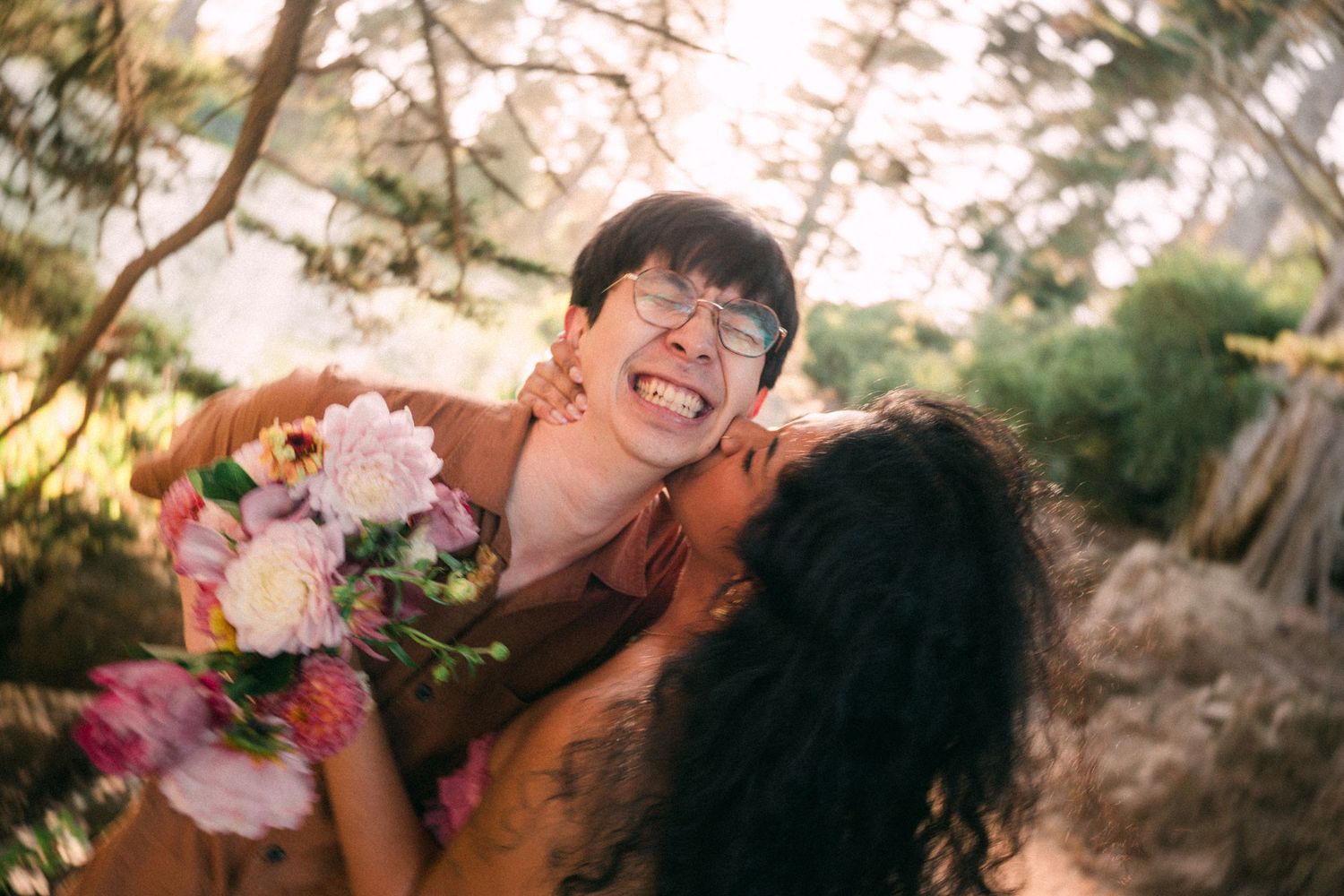 A joyful couple embraces outdoors, woman kissing mans cheek as he laughs holding a bouquet of pink flowers.