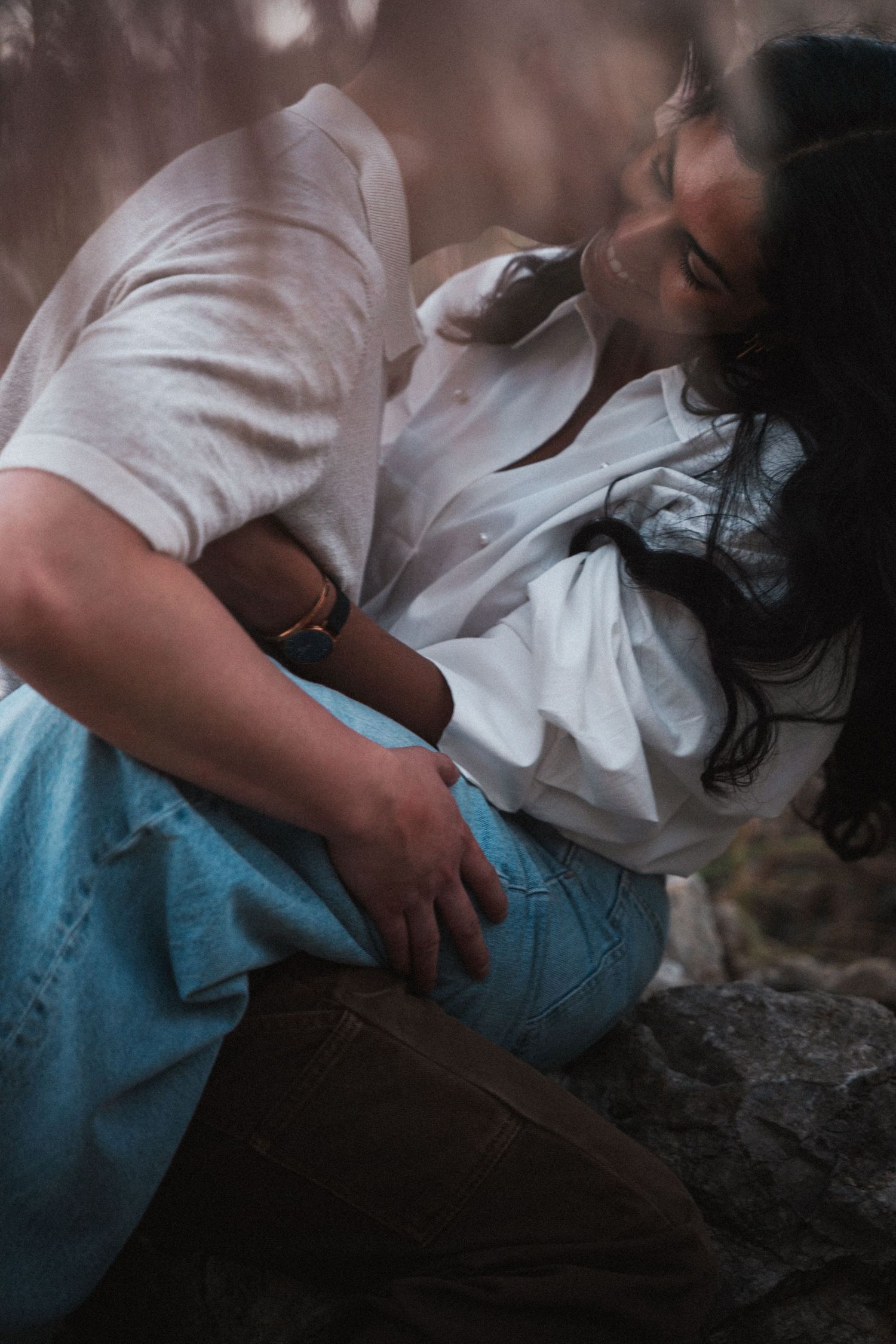 A couple sharing an intimate embrace outdoors, sitting on rocks in white shirts and casual attire.
