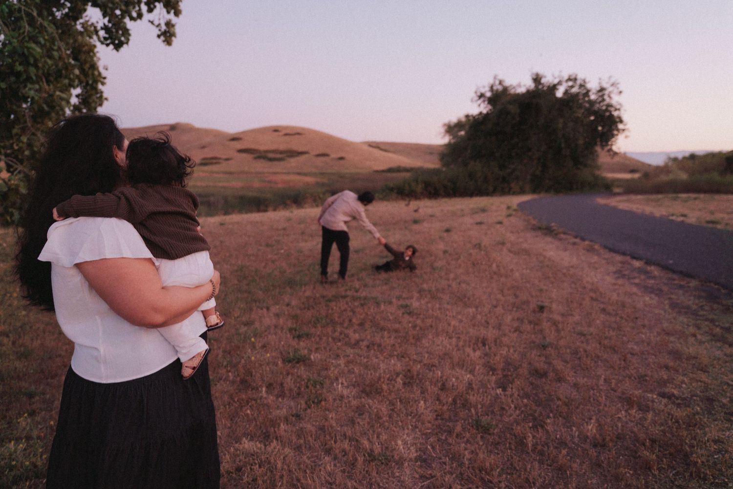 A woman holds a baby while watching a man play with a dog in a golden field at dusk near a winding road.