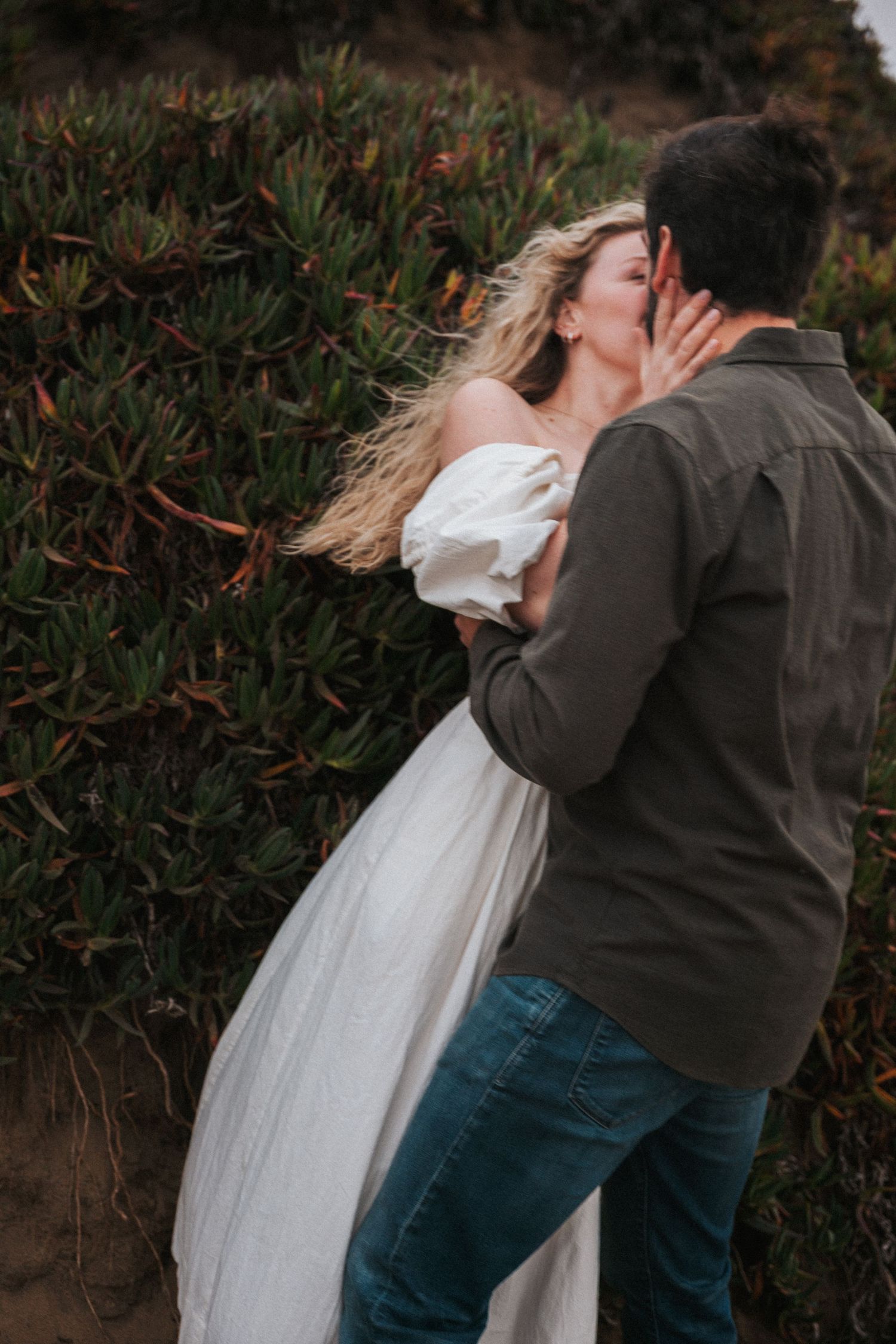 Couple sharing a romantic kiss outdoors near lush green bushes, woman in white dress with windswept hair.