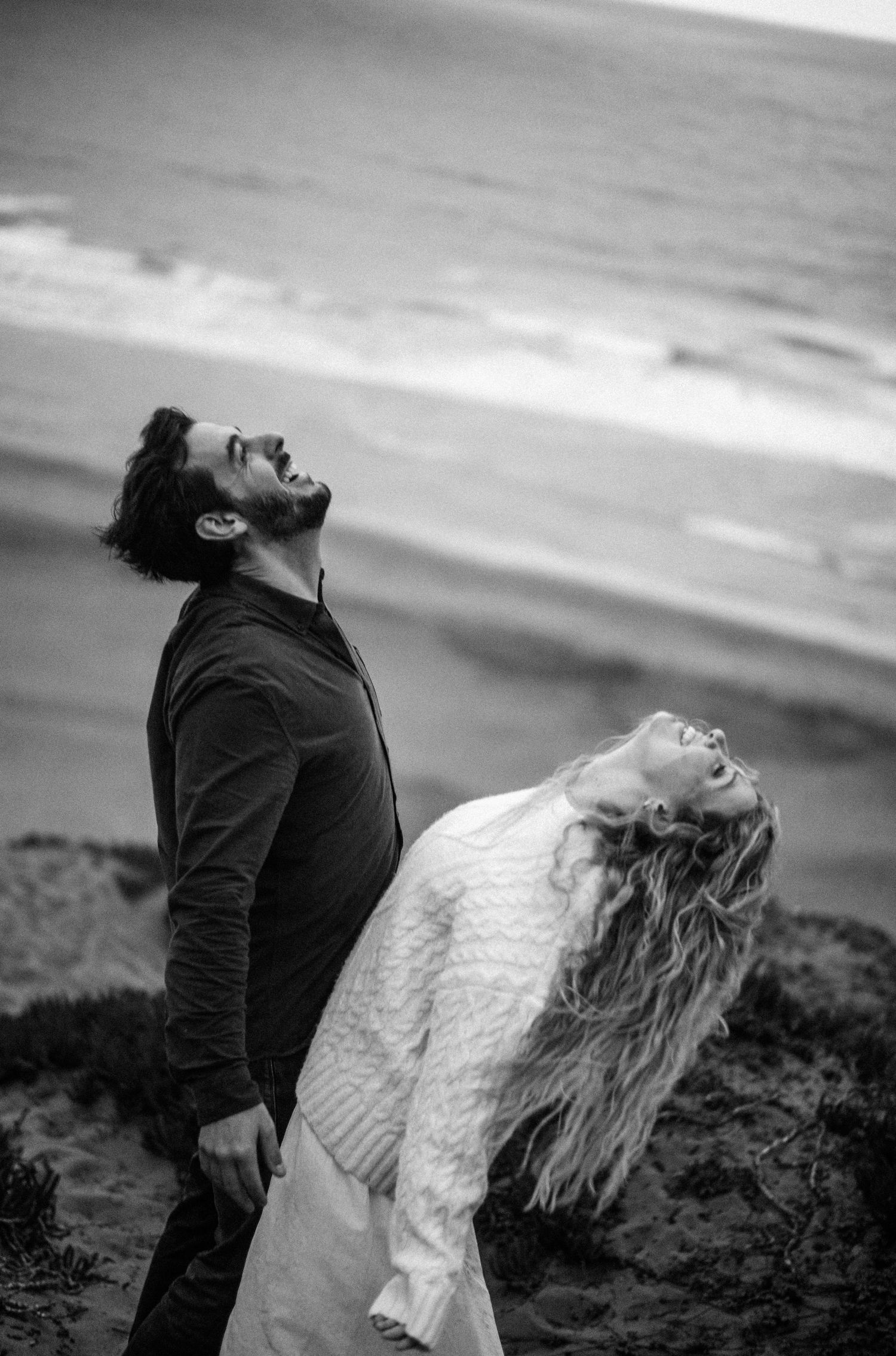 Black and white photo of a couple laughing with heads tilted back on a windy beach.