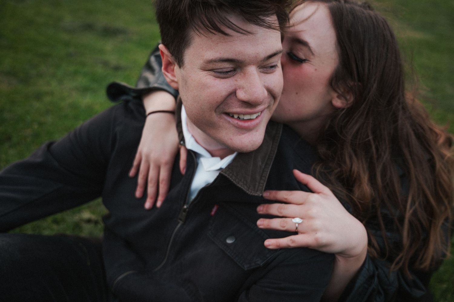 Happy couple sitting on grass, woman with engagement ring kissing smiling man on the cheek.