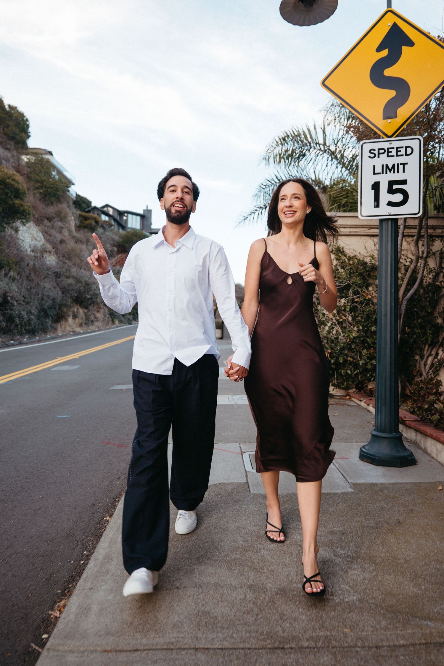 A smiling couple walks hand-in-hand on a sidewalk near a speed limit 15 sign on a sunny California street.