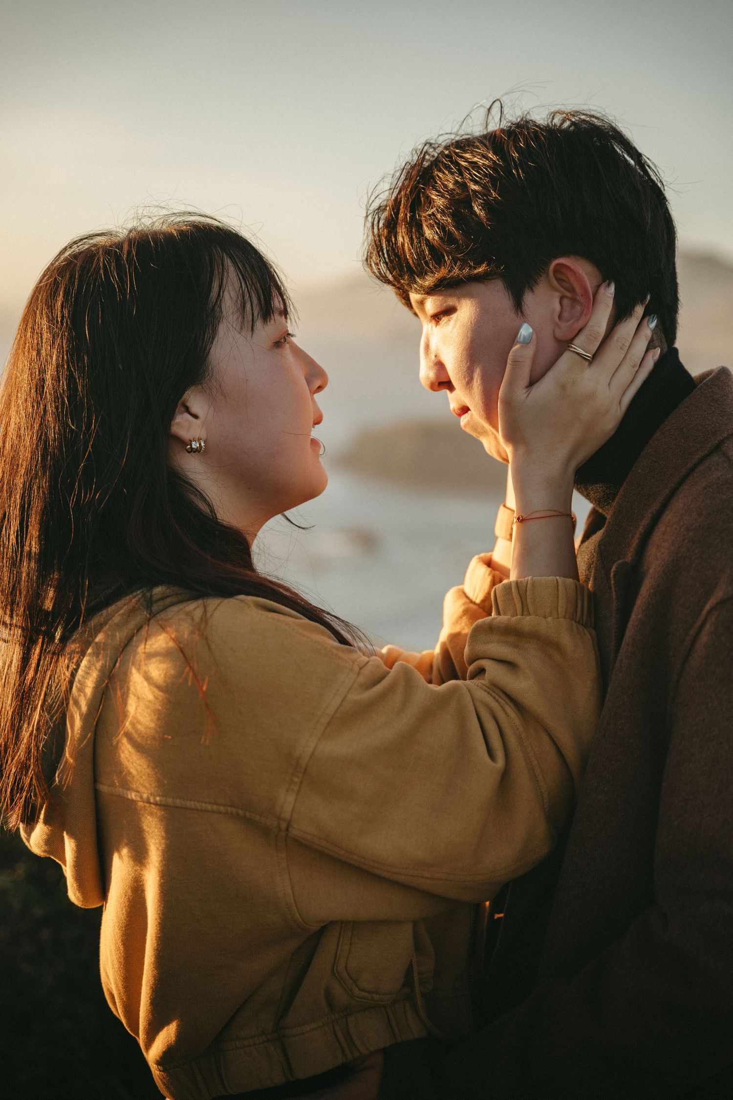 A couple shares a tender moment at sunset as a woman in a mustard jacket gently touches a man's face by the coast.