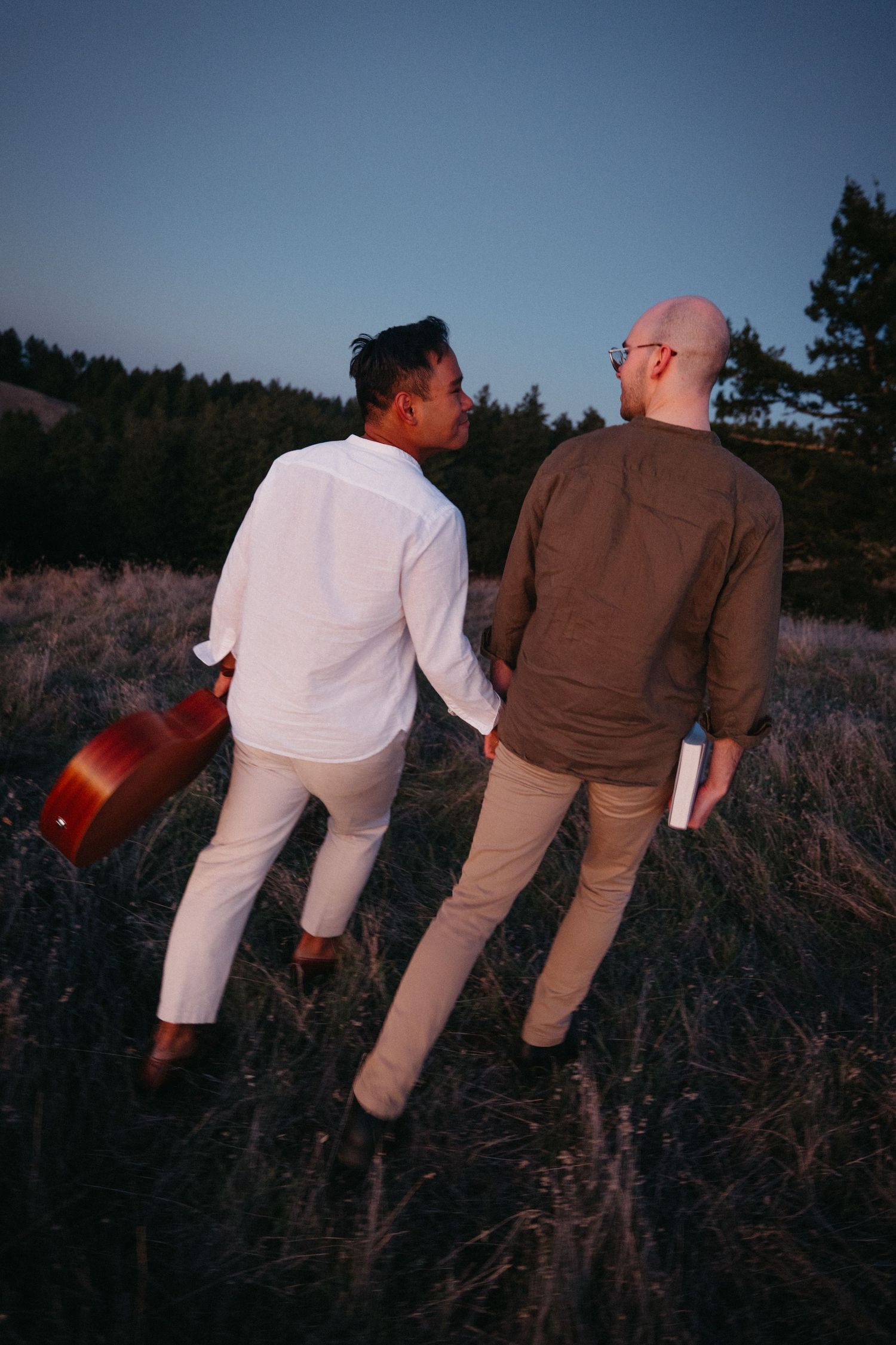 Two men walk through a field at dusk, one carrying a guitar and the other holding a book, enjoying nature.