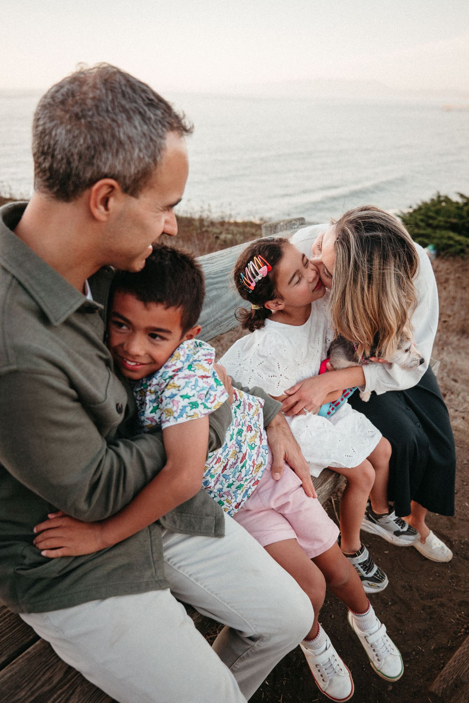 A happy family of four sits together outdoors near the ocean, with parents hugging their children warmly.