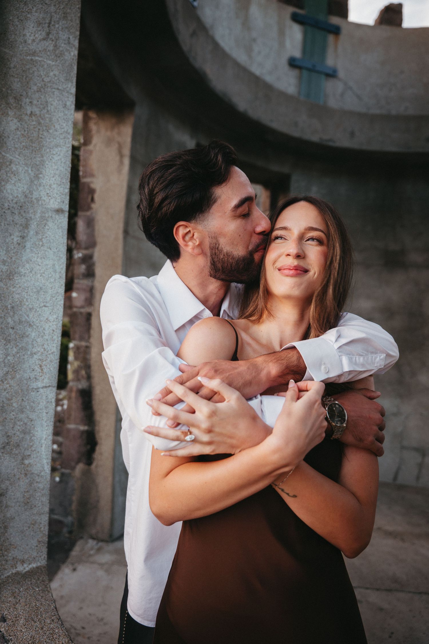 A man in a white shirt embraces and kisses a smiling woman in a black dress near stone columns.