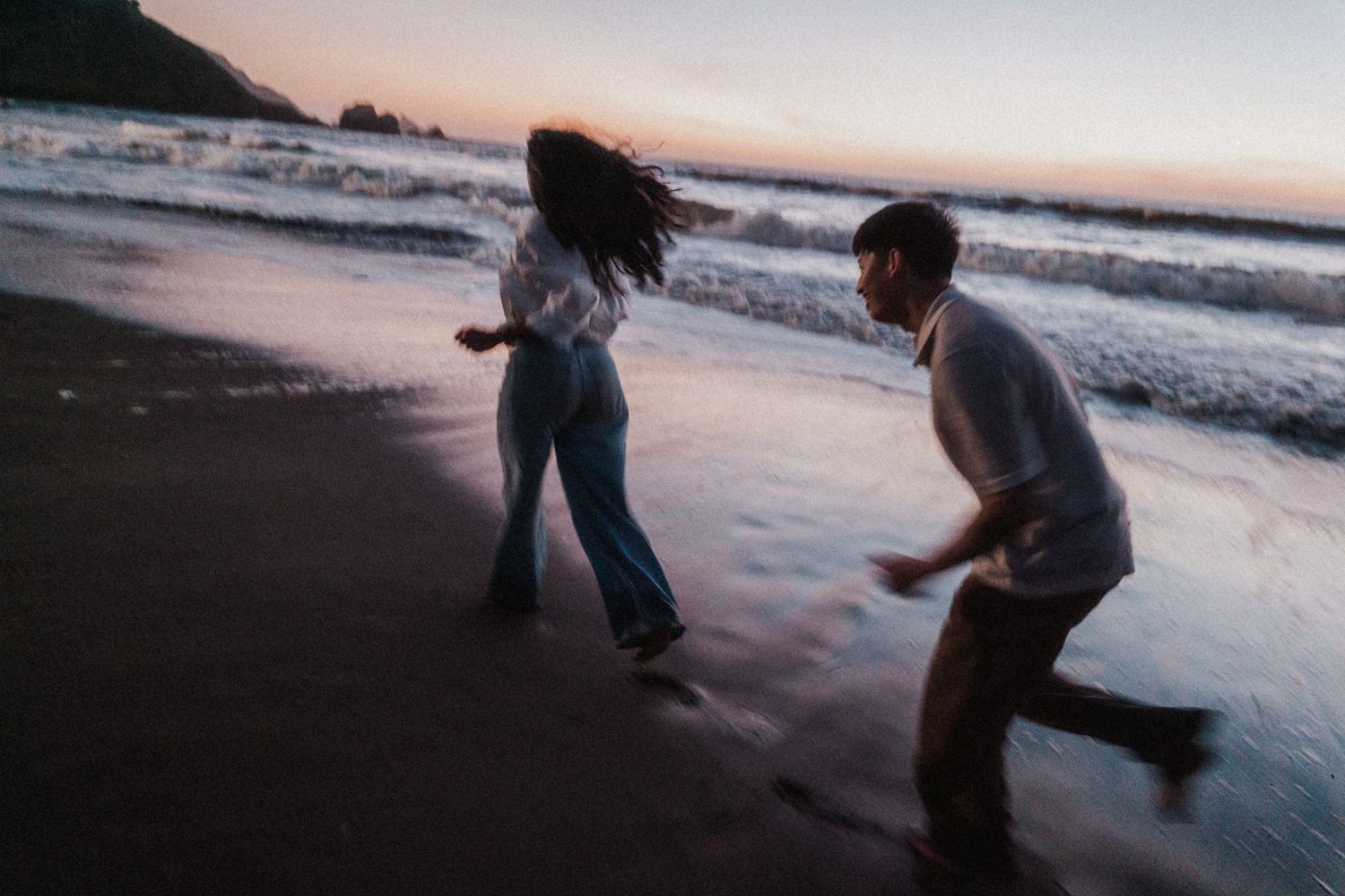 Couple playfully running on a dark sandy beach at sunset with ocean waves in the background.